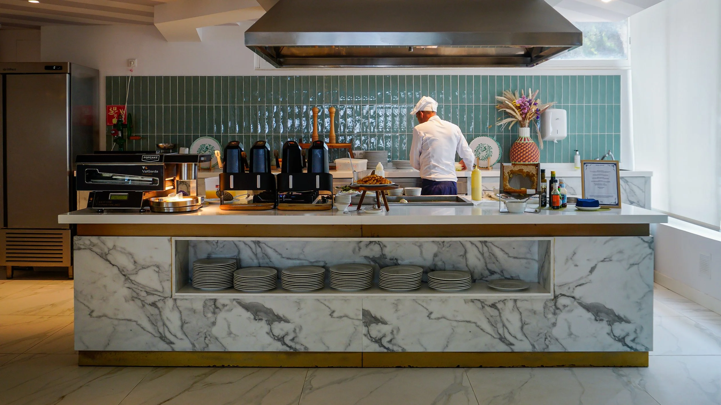 Breakfast buffet with a marble counter, green tiled walls, and a chef preparing fresh dishes in an open kitchen