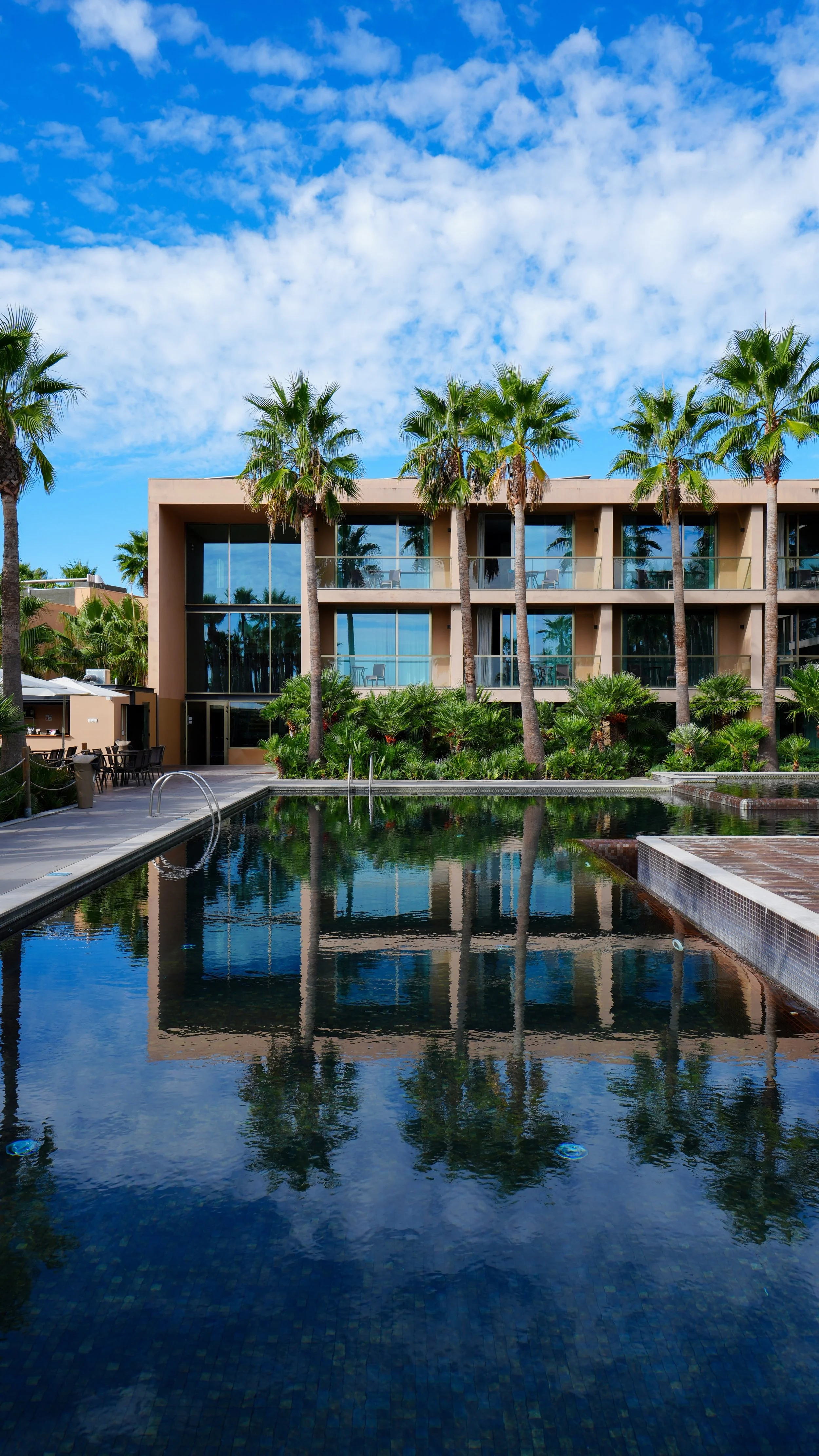 Modern resort building with glass balconies overlooking an outdoor swimming pool framed by palm trees