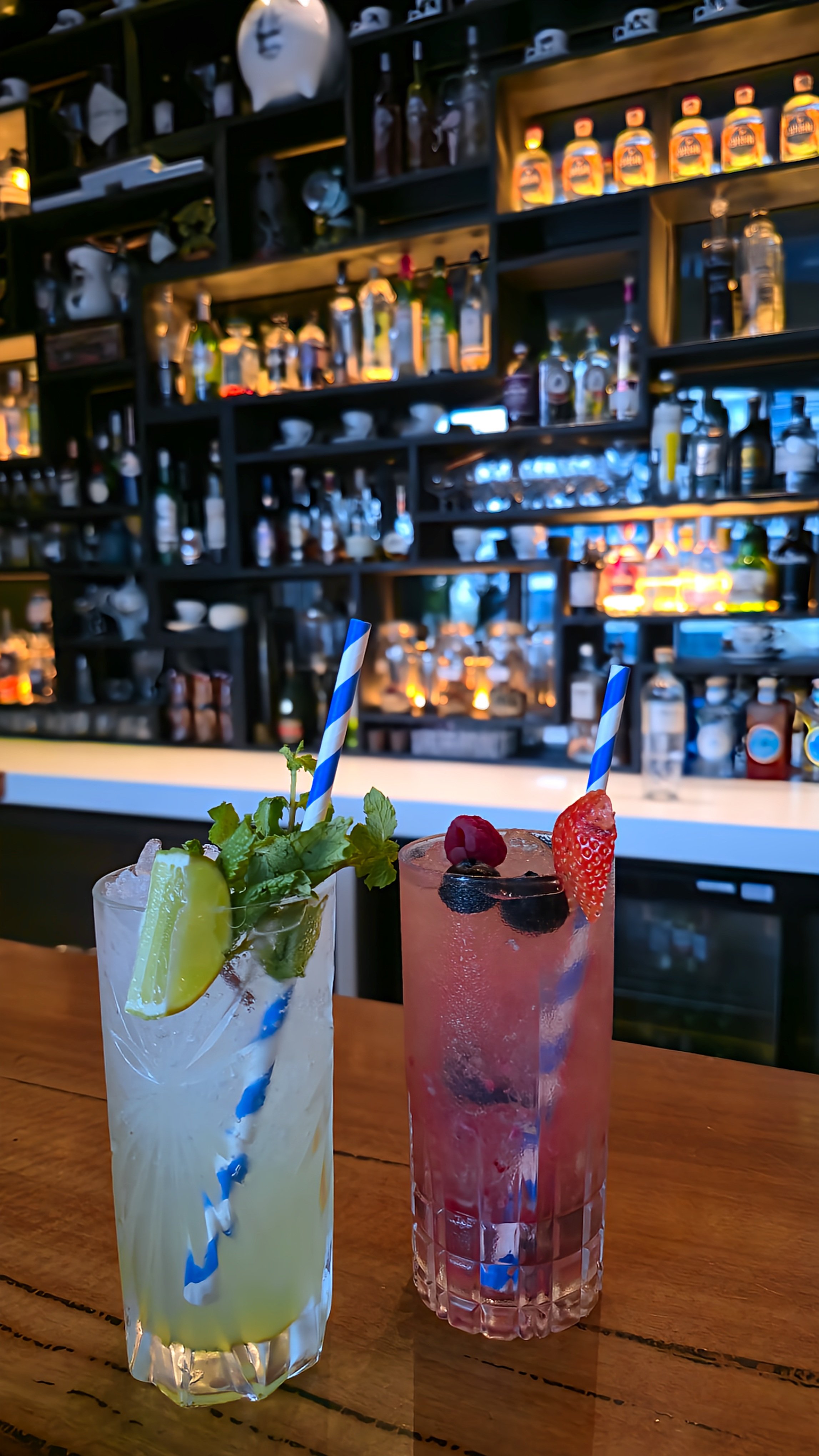 Two colorful cocktails garnished with fresh fruit and herbs against a backlit bar displaying an extensive spirits collection