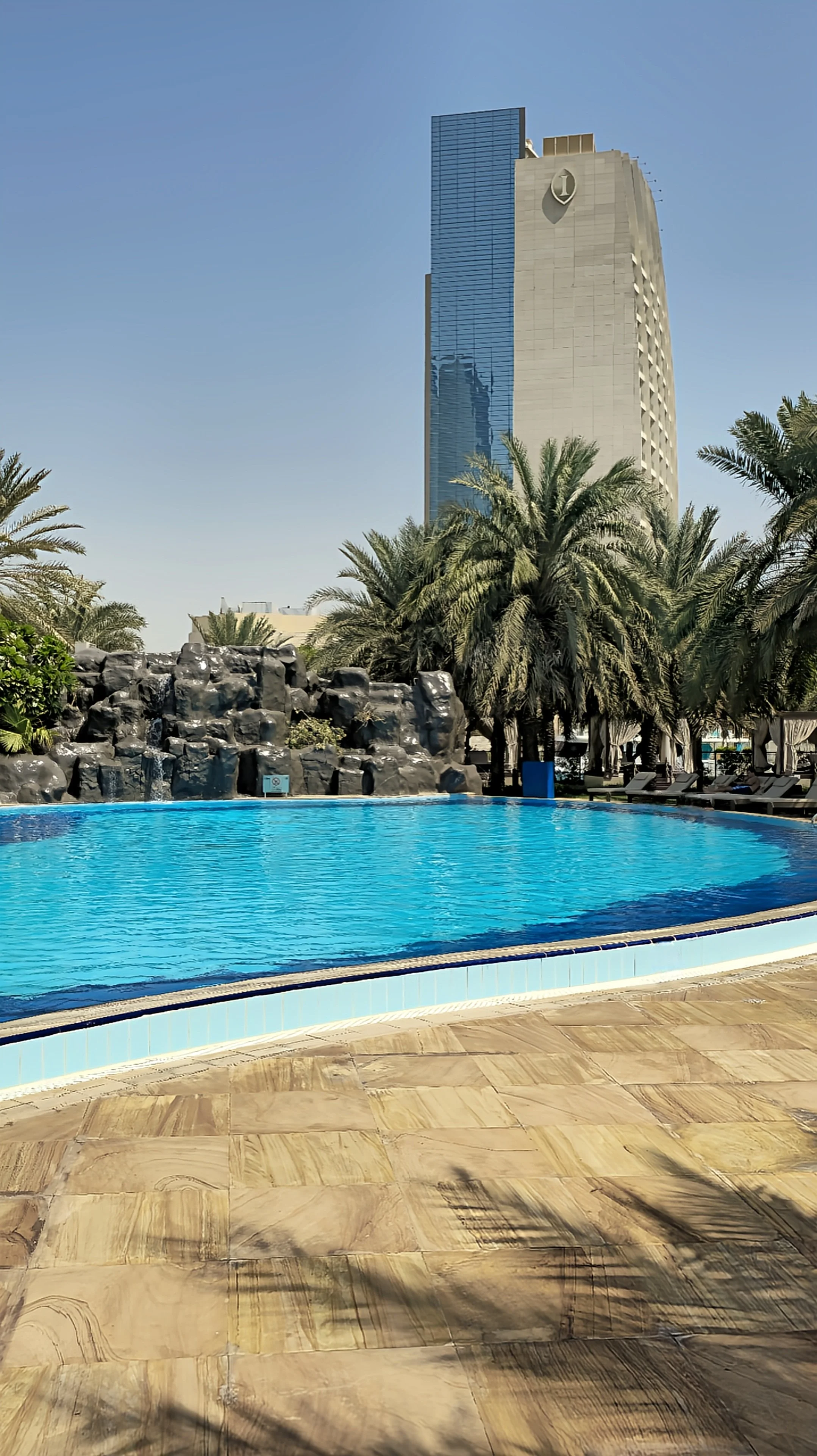 Outdoor swimming pool with turquoise water, artificial rock waterfall, palm trees, and a luxury hotel tower in the background