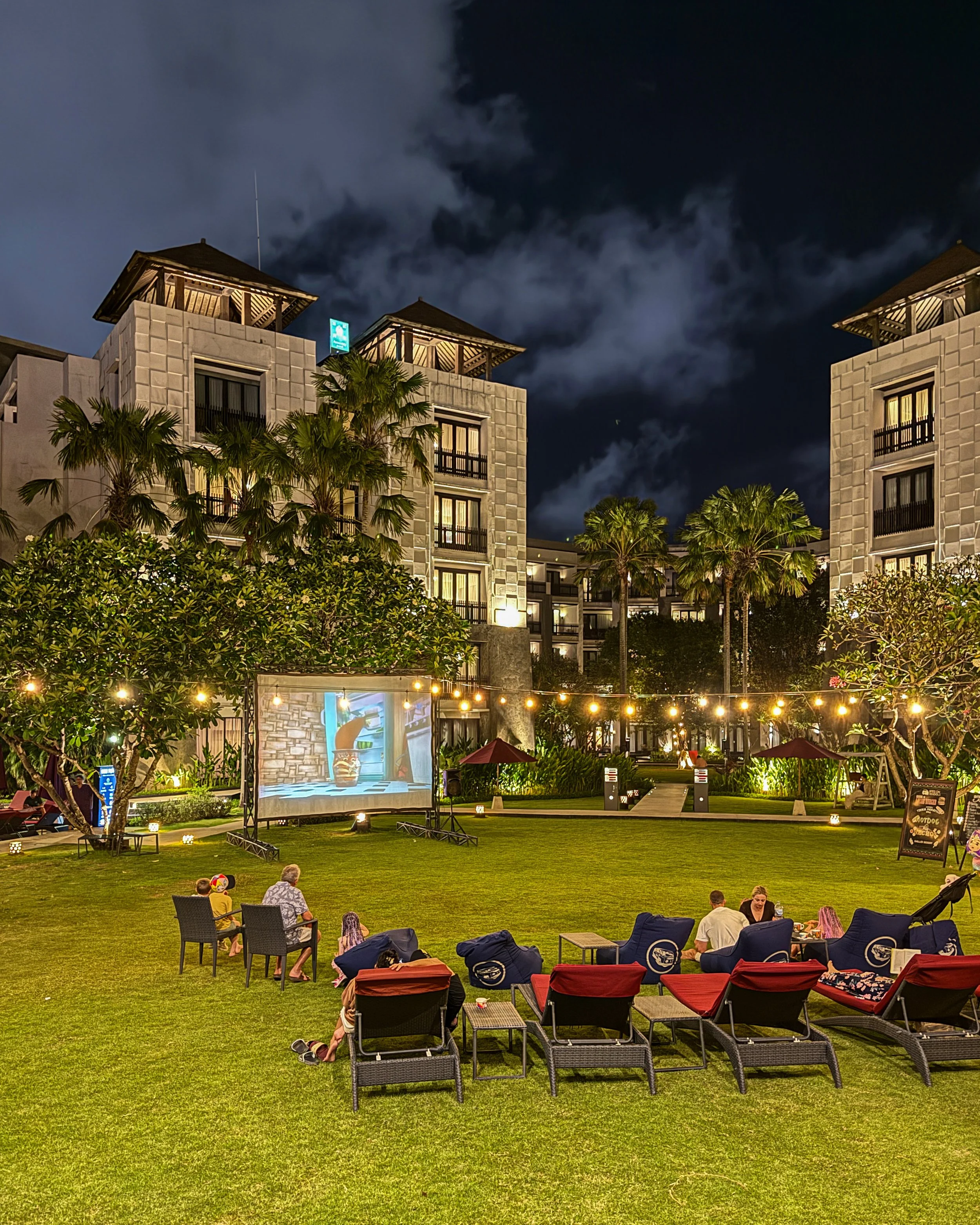 Guests enjoy an outdoor movie night on the garden lawn surrounded by illuminated palm trees and contemporary architecture