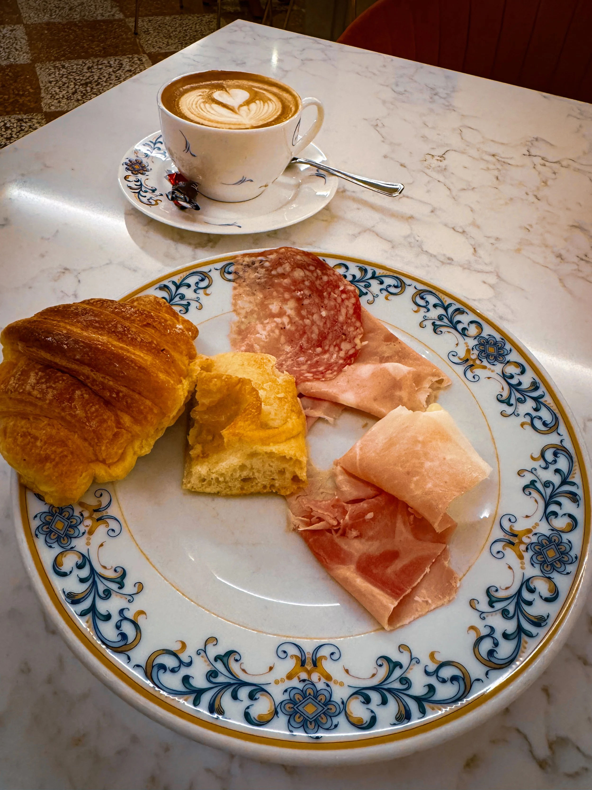 Italian breakfast with cappuccino, croissant, prosciutto, and salami on ceramic plate, Art Hotel Commercianti – Bologna, Italy.