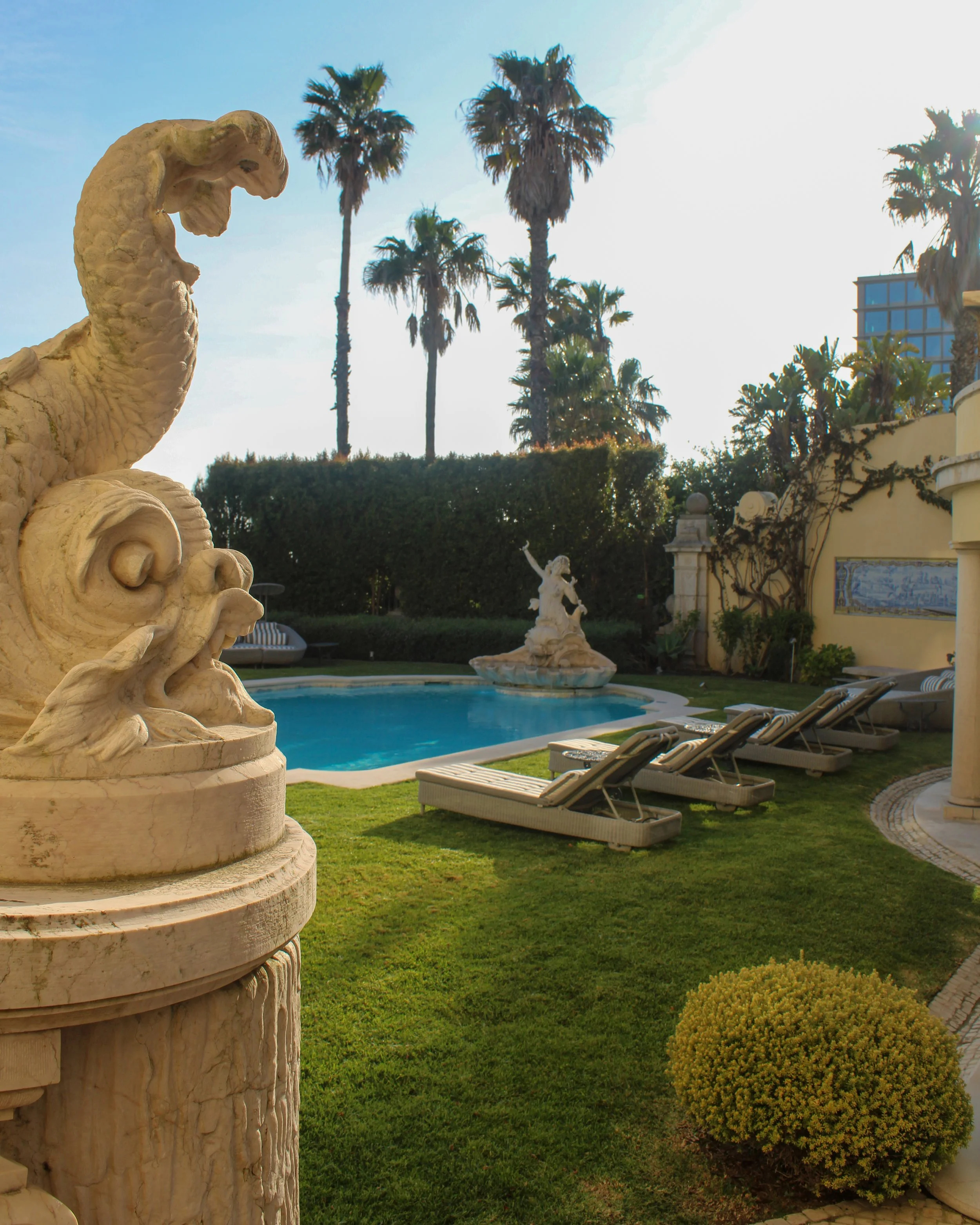 Poolside scene with sculptural fountain, palm trees, and sun loungers in a tranquil garden setting, Estoril Vintage Hotel – Estoril, Portugal.