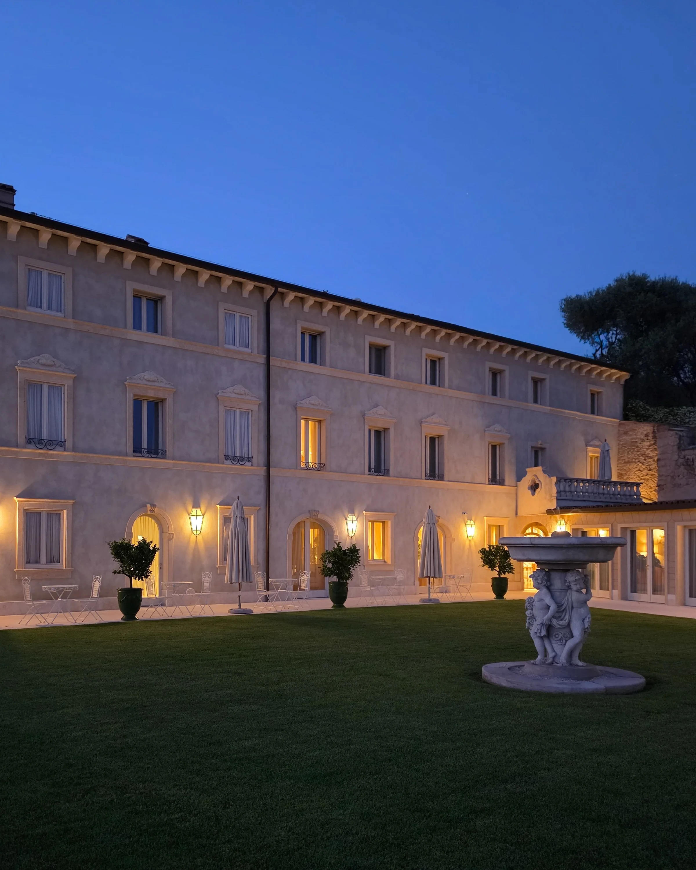 Elegant façade of Relais Fra’ Lorenzo illuminated at dusk with landscaped gardens and historic fountain