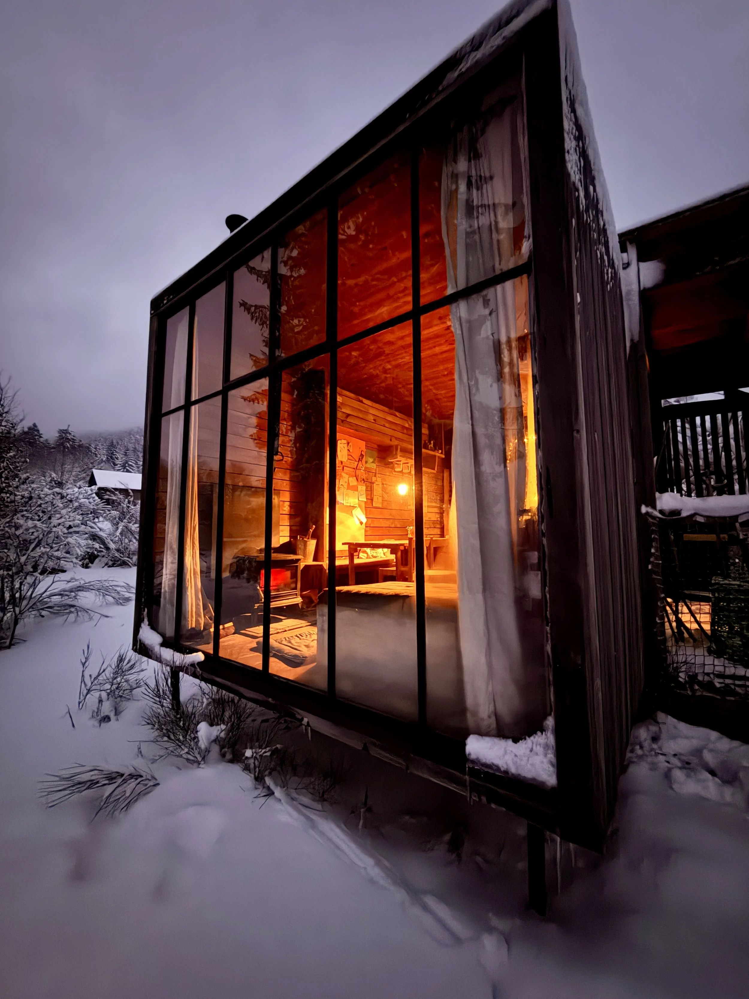 Modern glass-fronted cabin glowing warmly against the snowy forest landscape at dusk