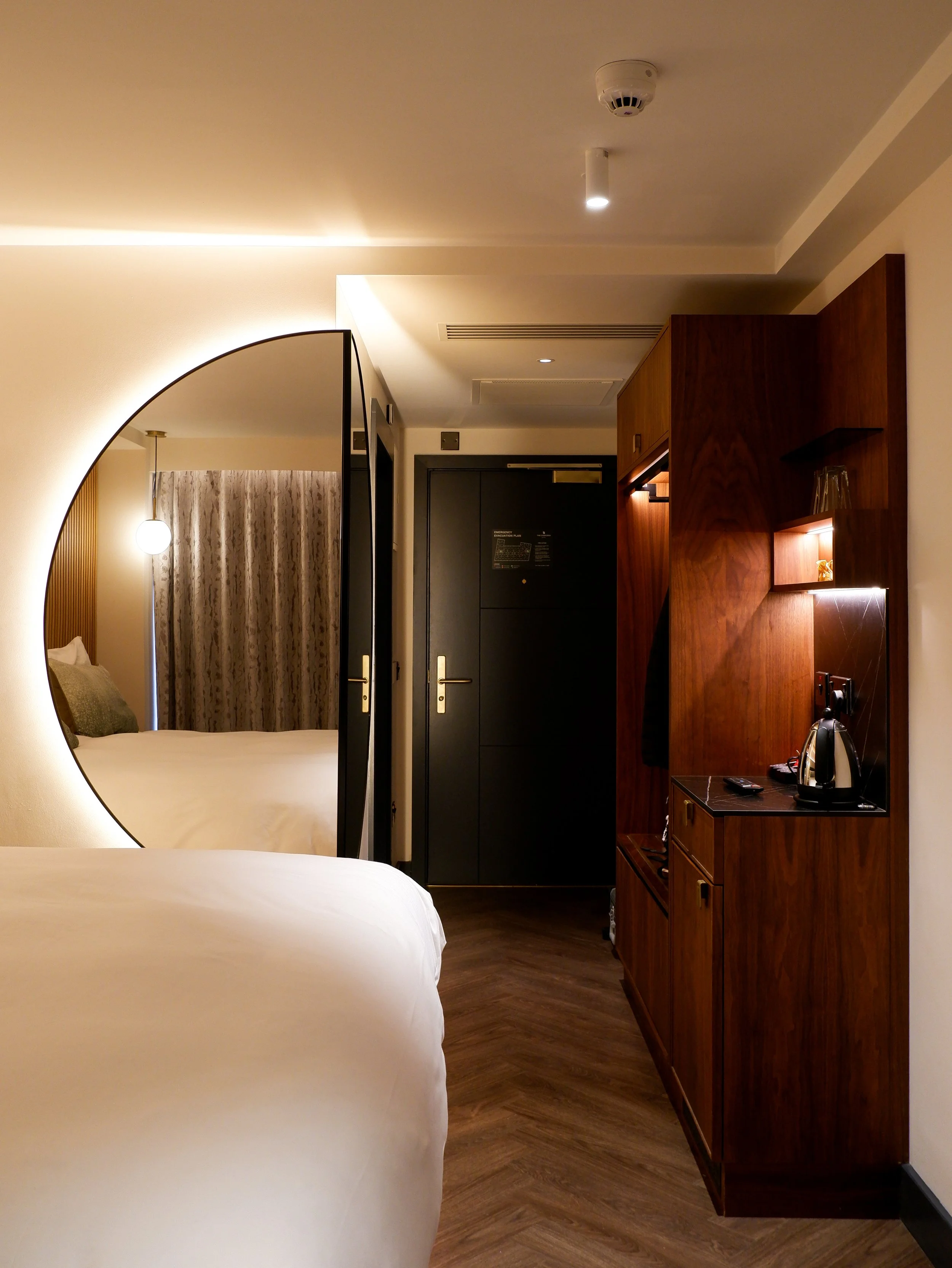 Contemporary guest room with warm wooden furnishings, a large illuminated mirror, and a coffee station