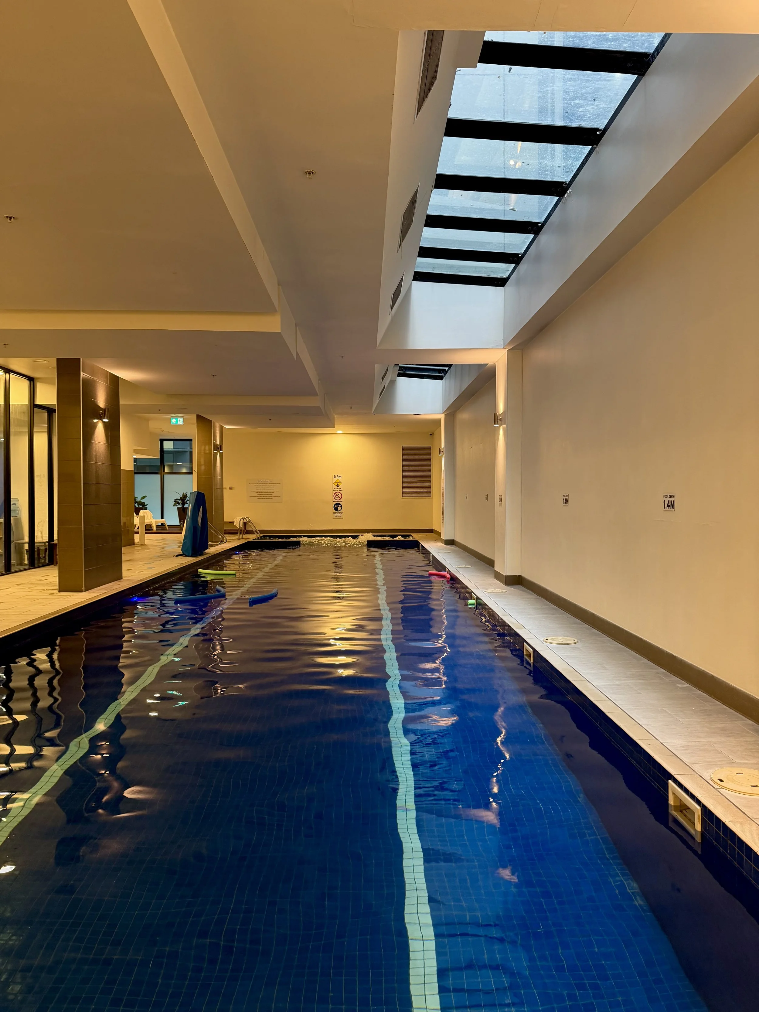 Indoor heated swimming pool with blue tiled lanes, warm lighting, and a glass skylight ceiling