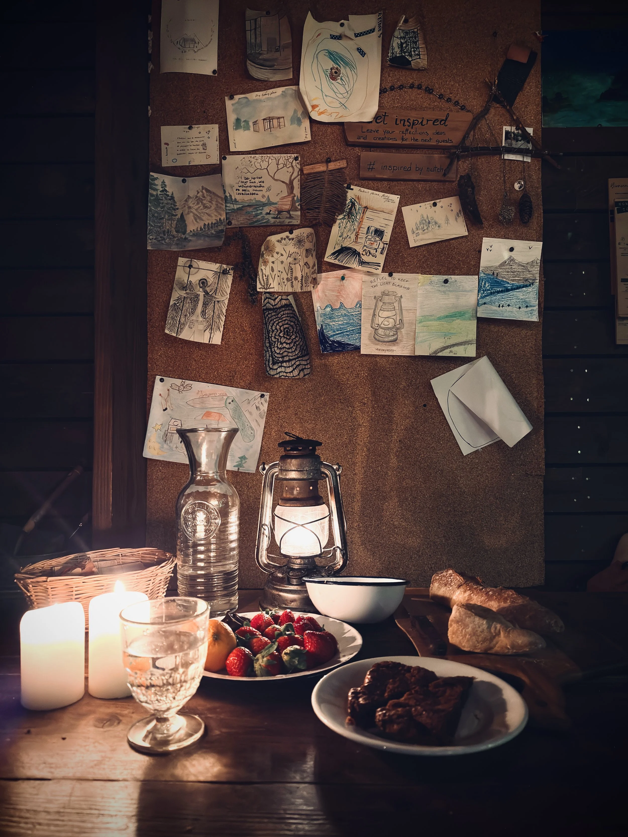 A warm, candlelit table set with simple breakfast dishes, fresh fruit, and handwritten sketches pinned on wood, creating a slow and intimate cabin atmosphere, Nutchel Forest Village Alsace – Plaine, France.
