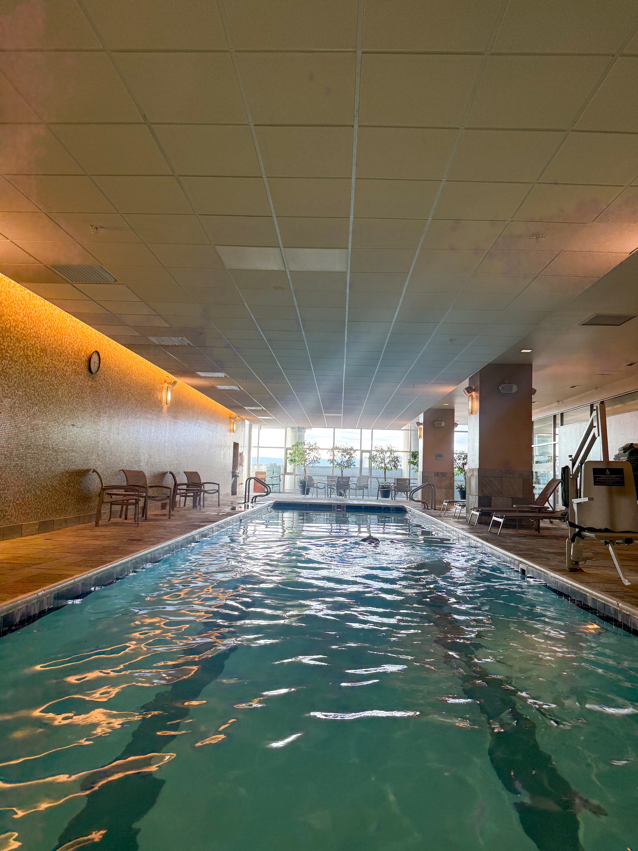 Indoor swimming pool with warm ambient lighting, comfortable seating, and large windows overlooking the city