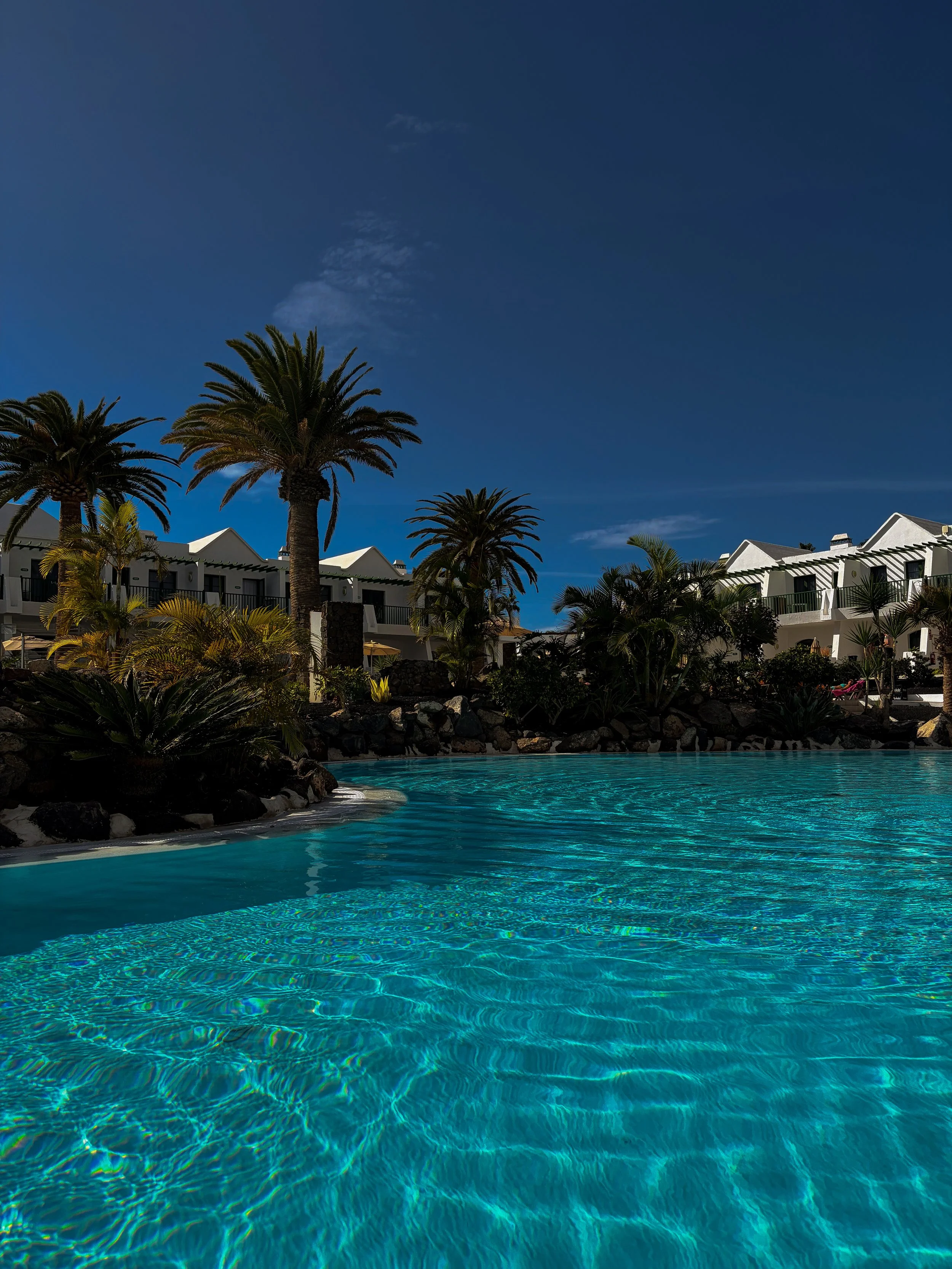 Outdoor swimming pool with turquoise water surrounded by palm trees, tropical gardens, and whitewashed hotel buildings