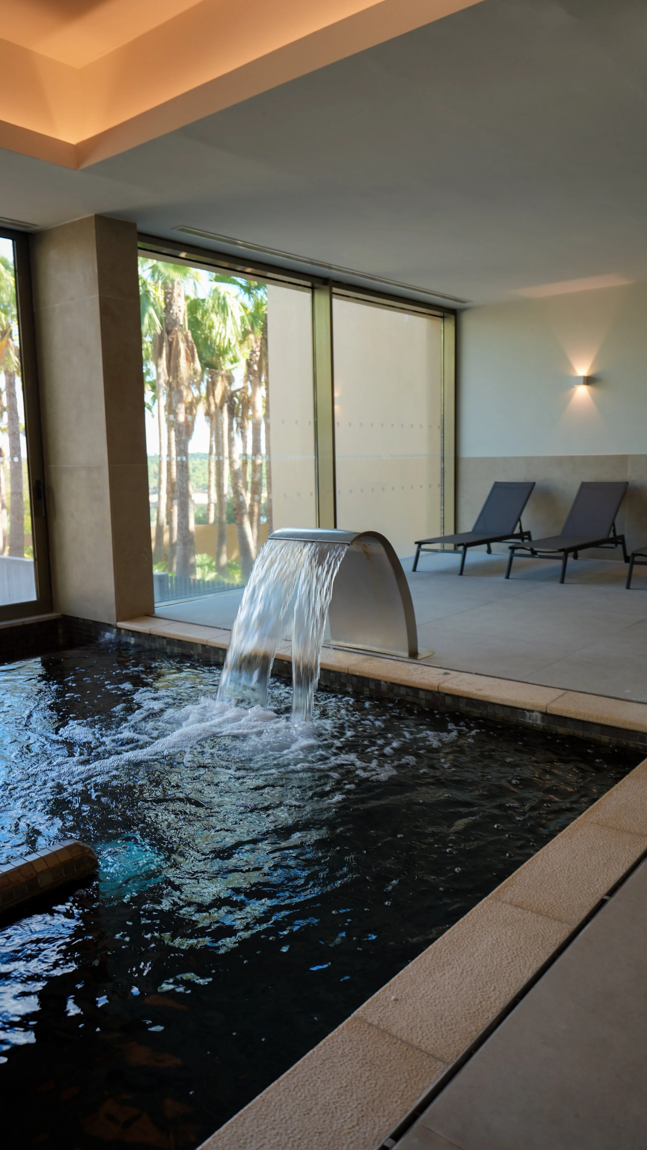 Indoor spa pool with a cascading water feature, relaxation loungers, and views of tropical palm trees