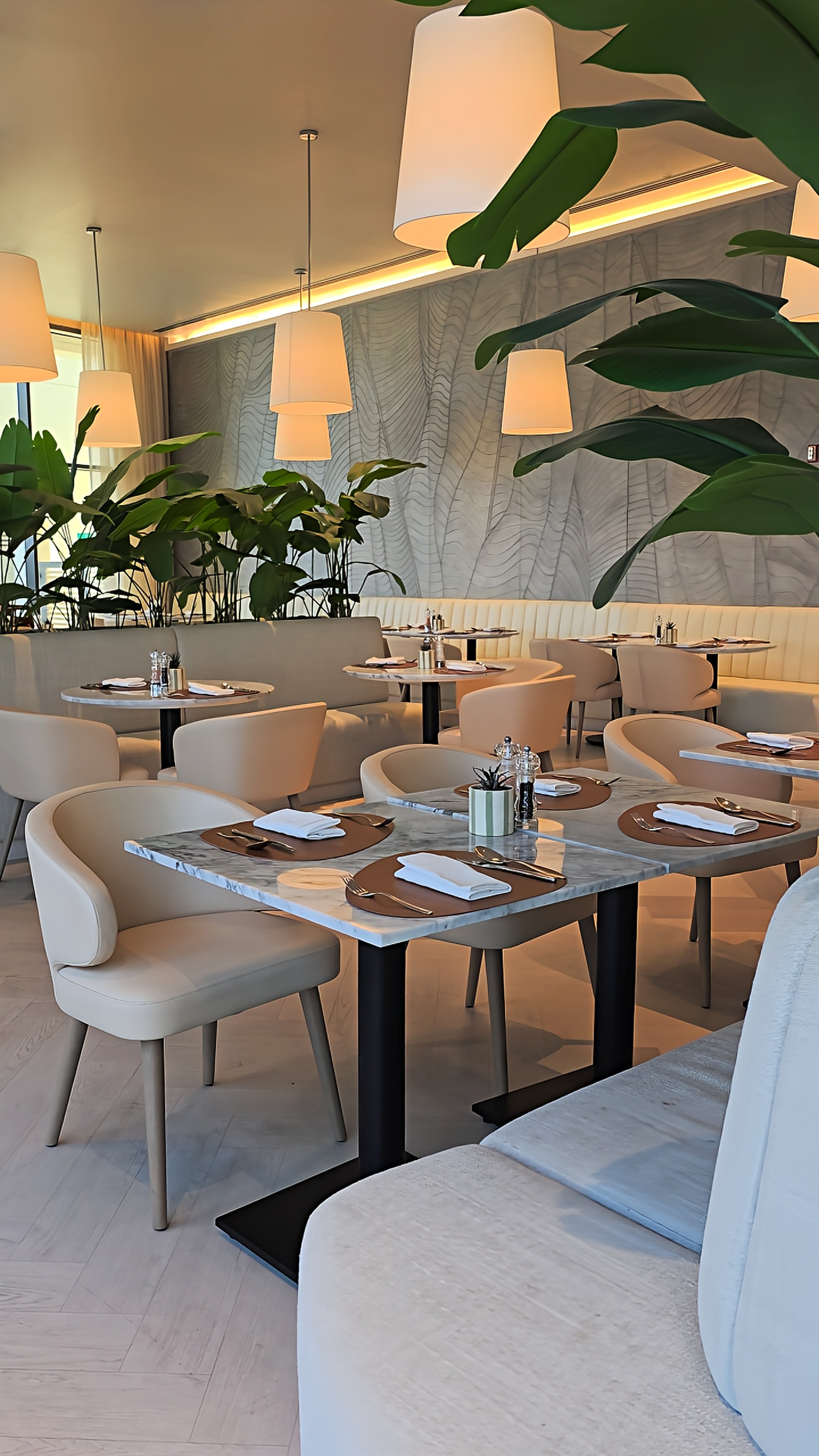 Modern restaurant interior featuring marble dining tables, cream upholstered chairs, tropical plants, and pendant lighting