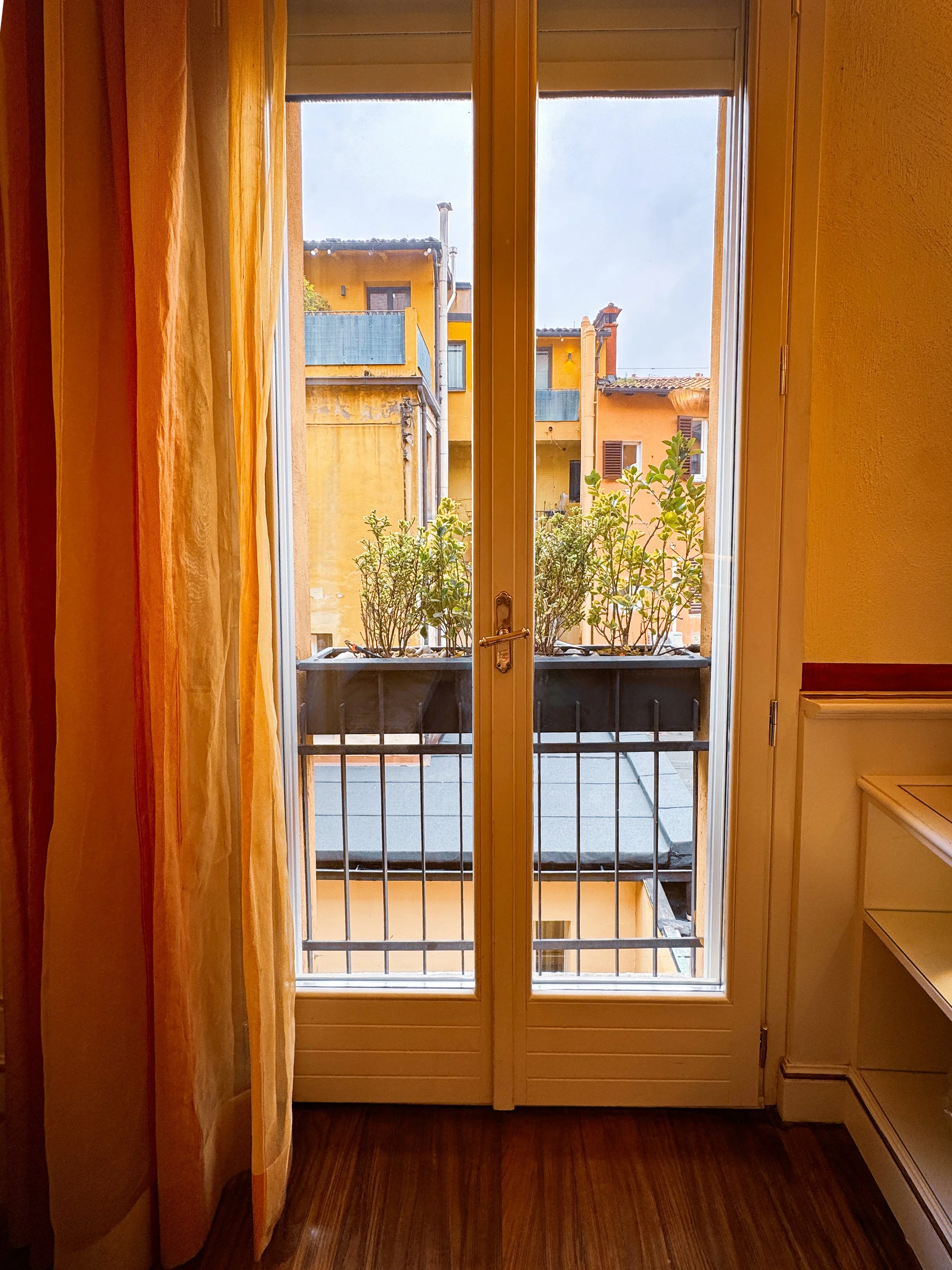 Balcony view over historic Bologna rooftops framed by tall French doors, Art Hotel Commercianti – Bologna, Italy.