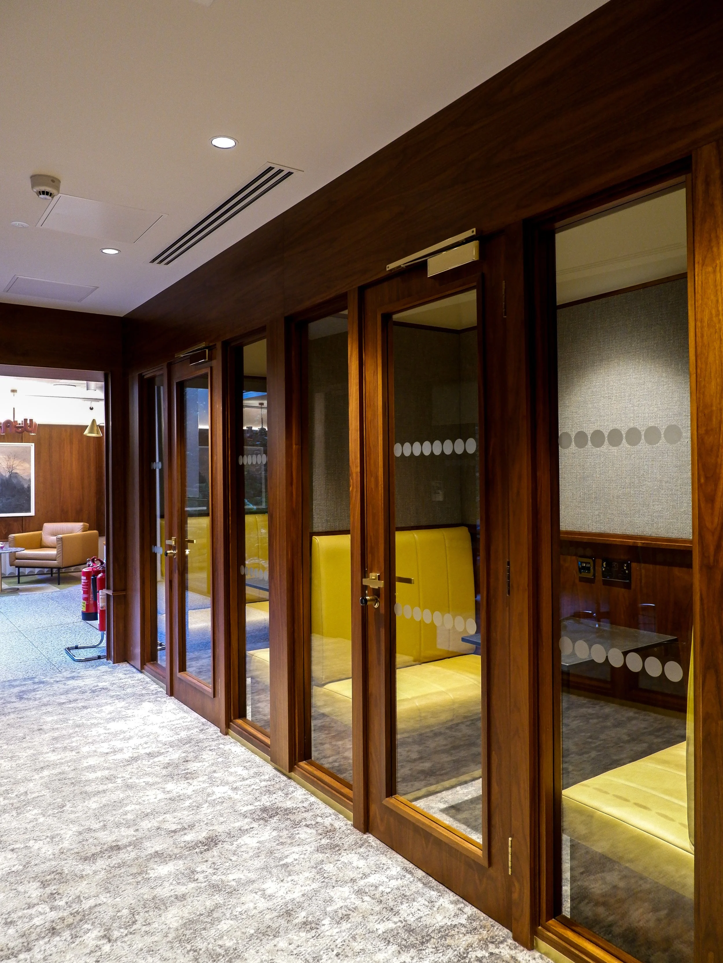 Private meeting booths with glass doors, warm wooden finishes, and yellow seating in a contemporary hotel workspace