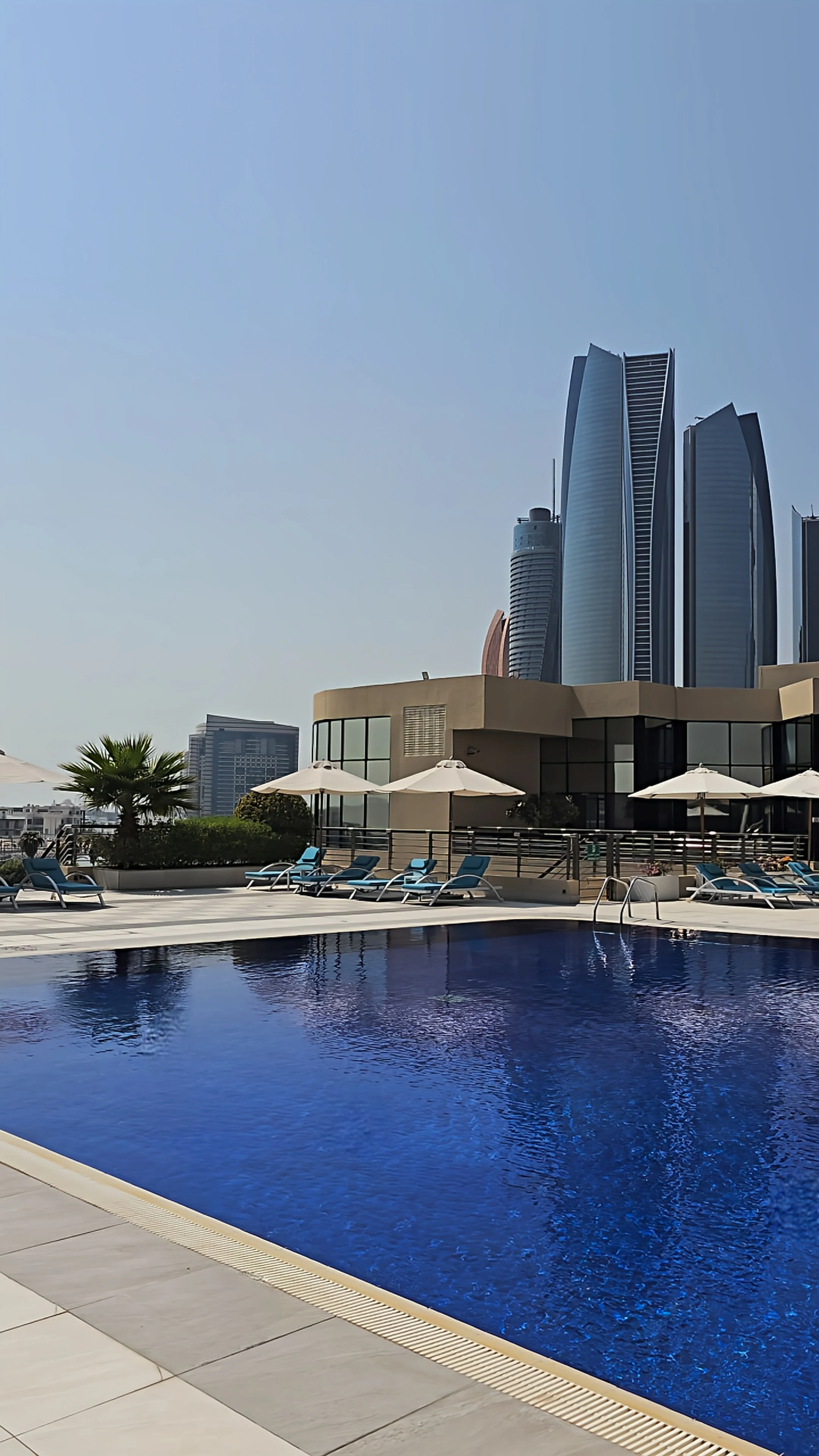 Rooftop swimming pool with blue water, sun loungers, white umbrellas, and modern skyscrapers overlooking Abu Dhabi
