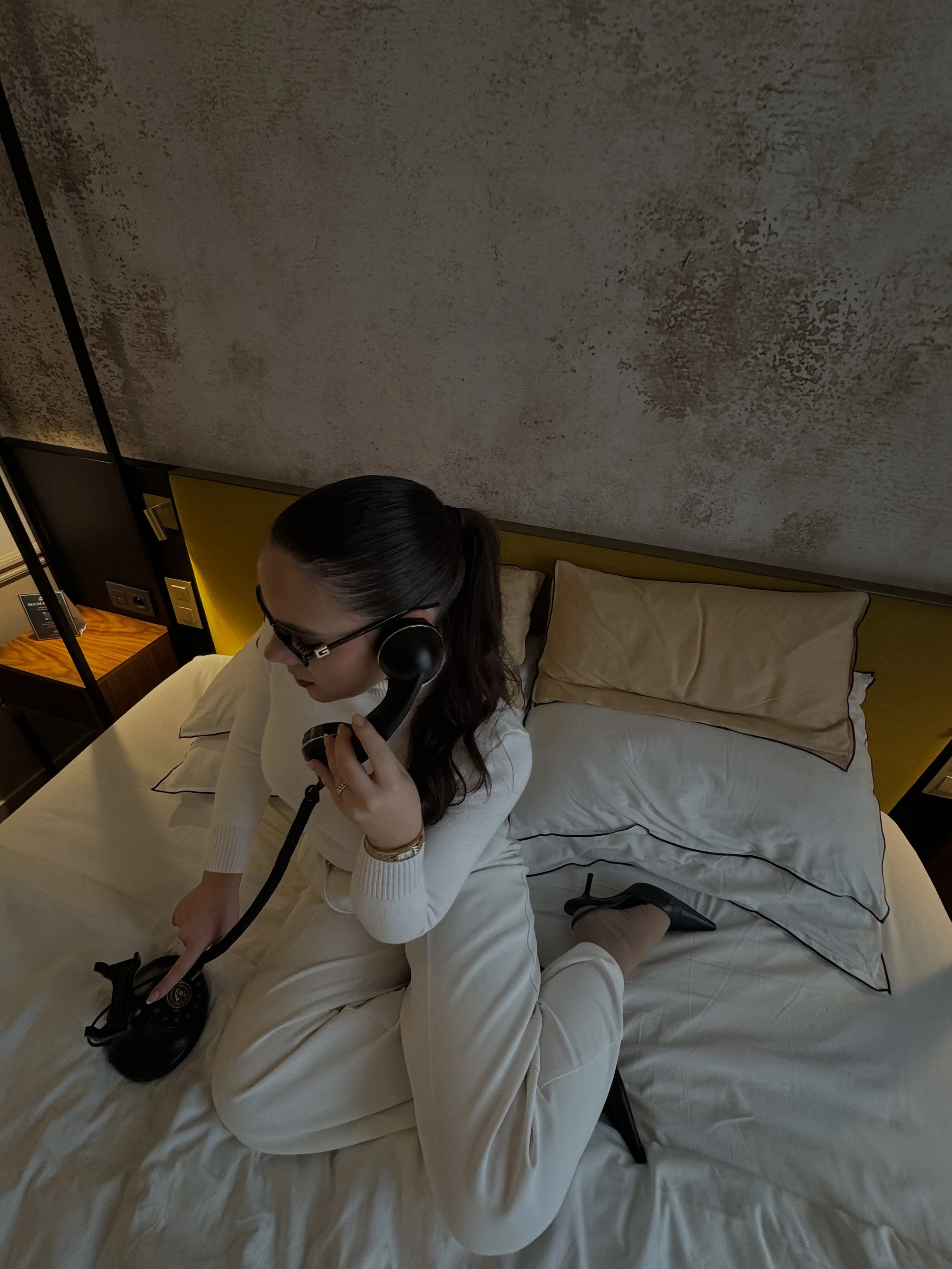 Stylish guest seated on a bed using a vintage telephone in a warmly lit modern-classic suite, Victoria Palace Hotel – Paris, France.
