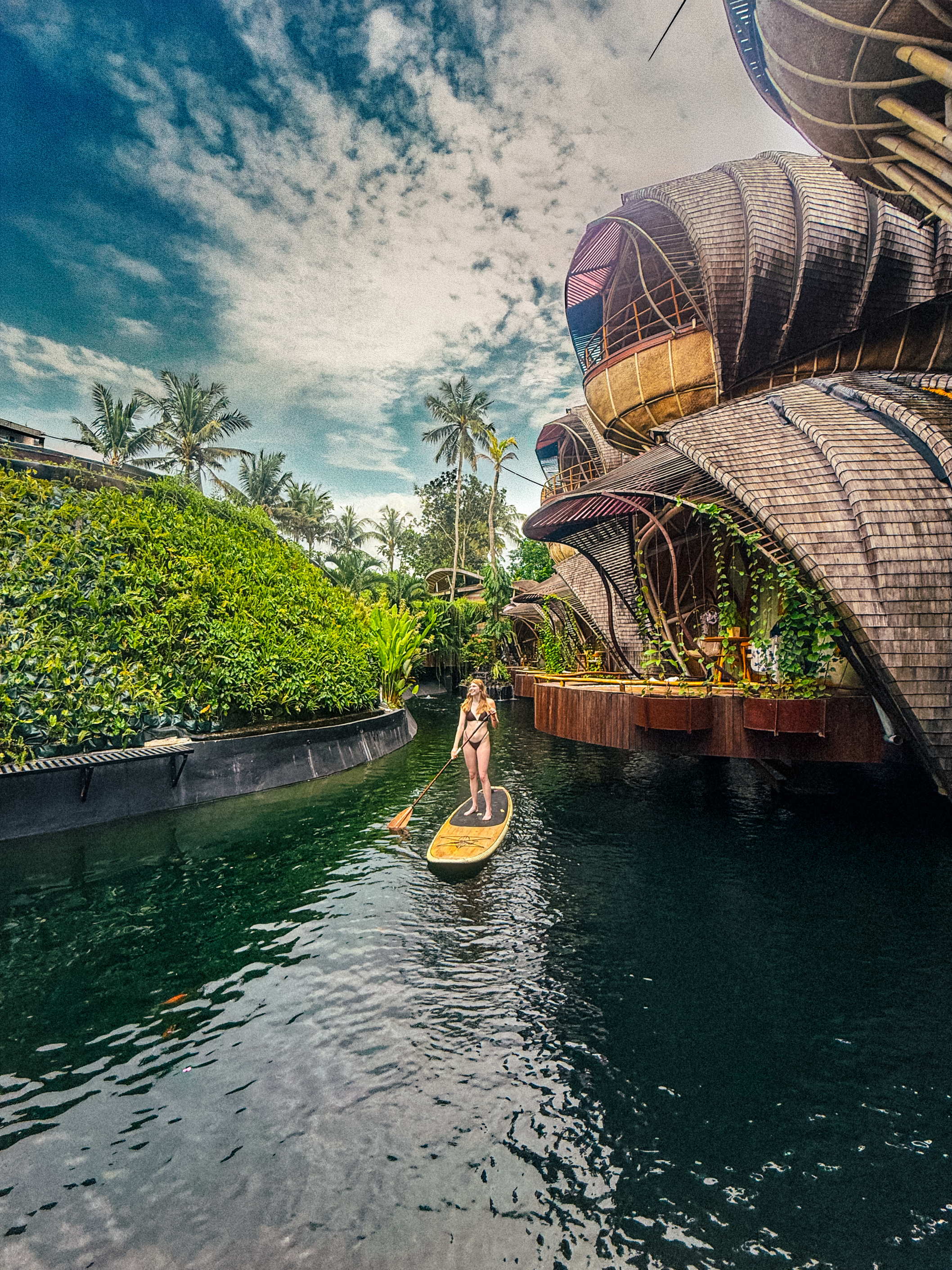 A guest paddleboards through tranquil waterways framed by iconic bamboo architecture and lush tropical gardens at this eco-inspired luxury resort in Bali