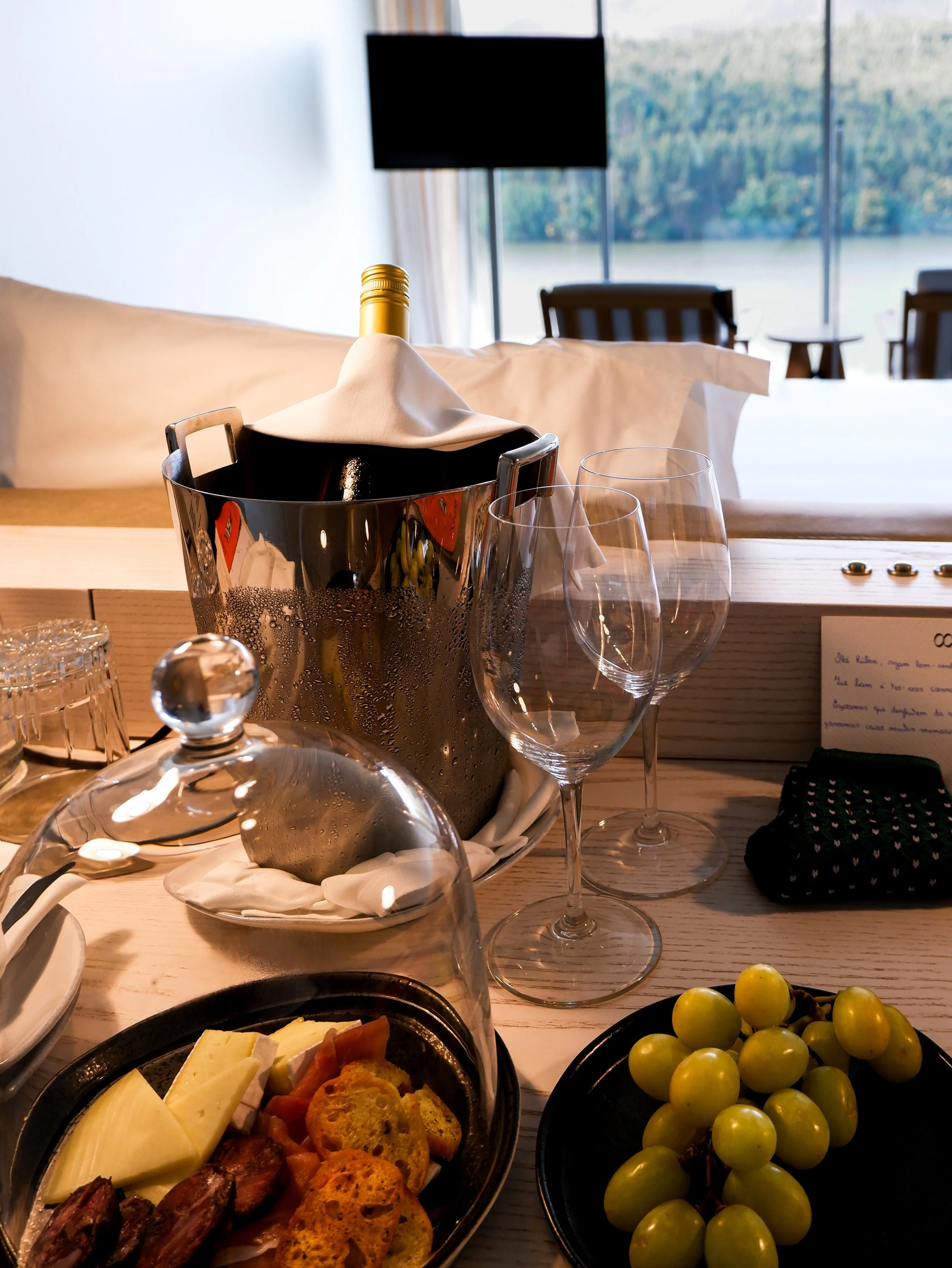 Luxury in-room dining setup with wine, cheese platter, and river views through floor-to-ceiling windows, offering a refined and relaxing stay, Octant Douro – Castelo de Paiva, Portugal.