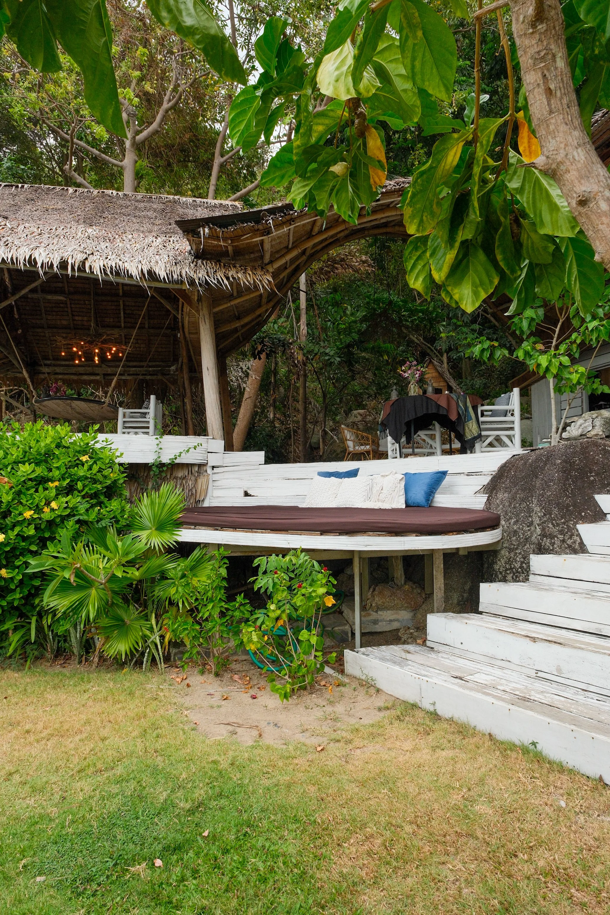 Open-air tropical pavilion with thatched architecture, cushioned seating, white timber, and lush greenery