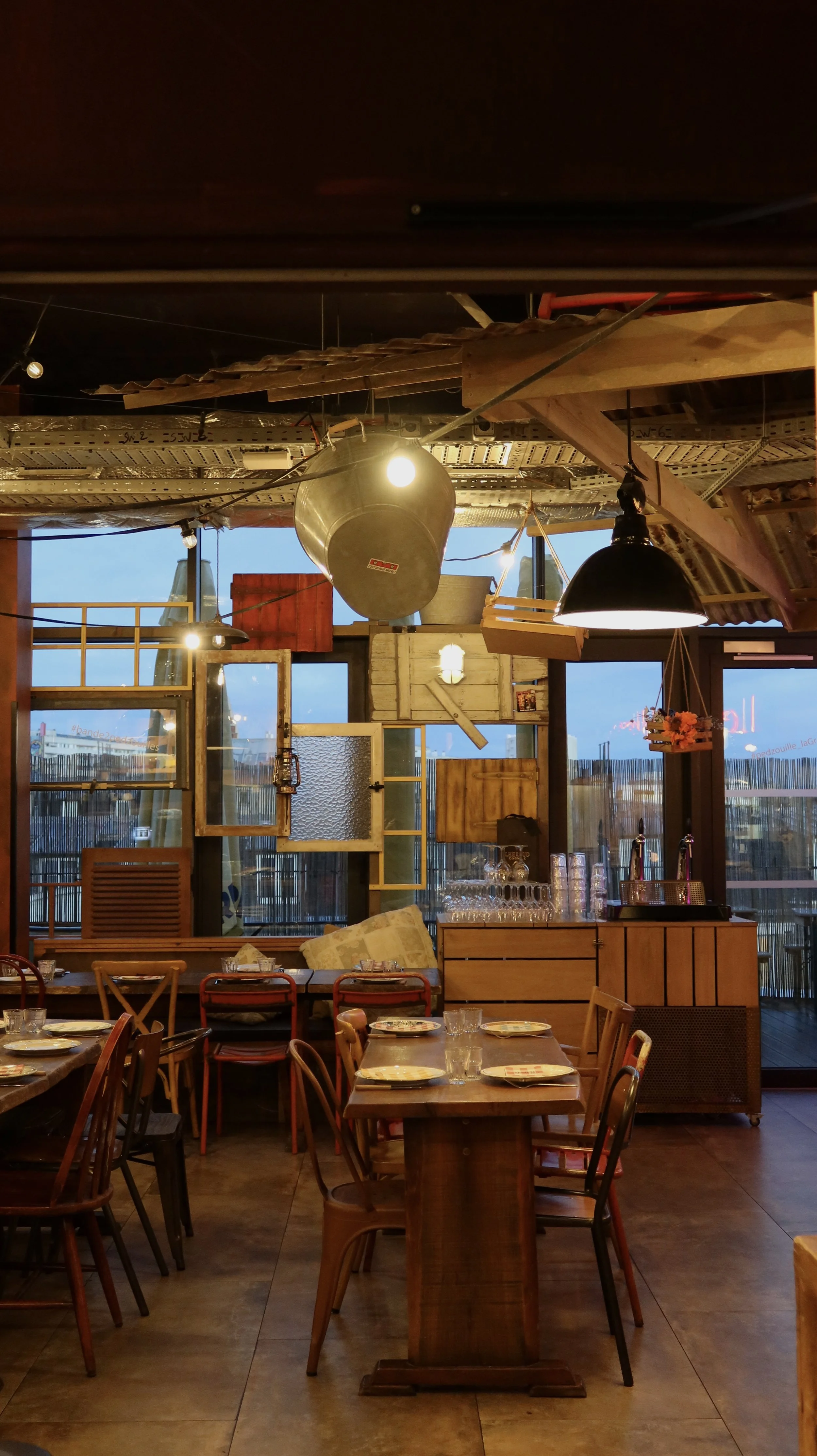 Warm industrial-style restaurant interior with wooden tables, open windows, and relaxed evening ambiance, Novotel Paris Porte de Versailles – Paris, France.