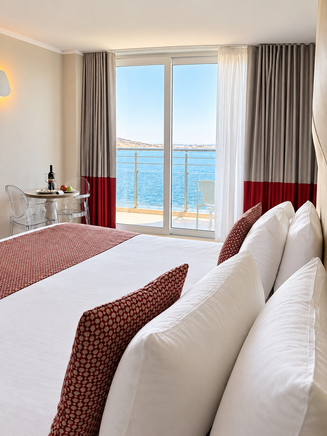 Bright sea-view room with crisp white bedding, red accents, and a private balcony overlooking the water