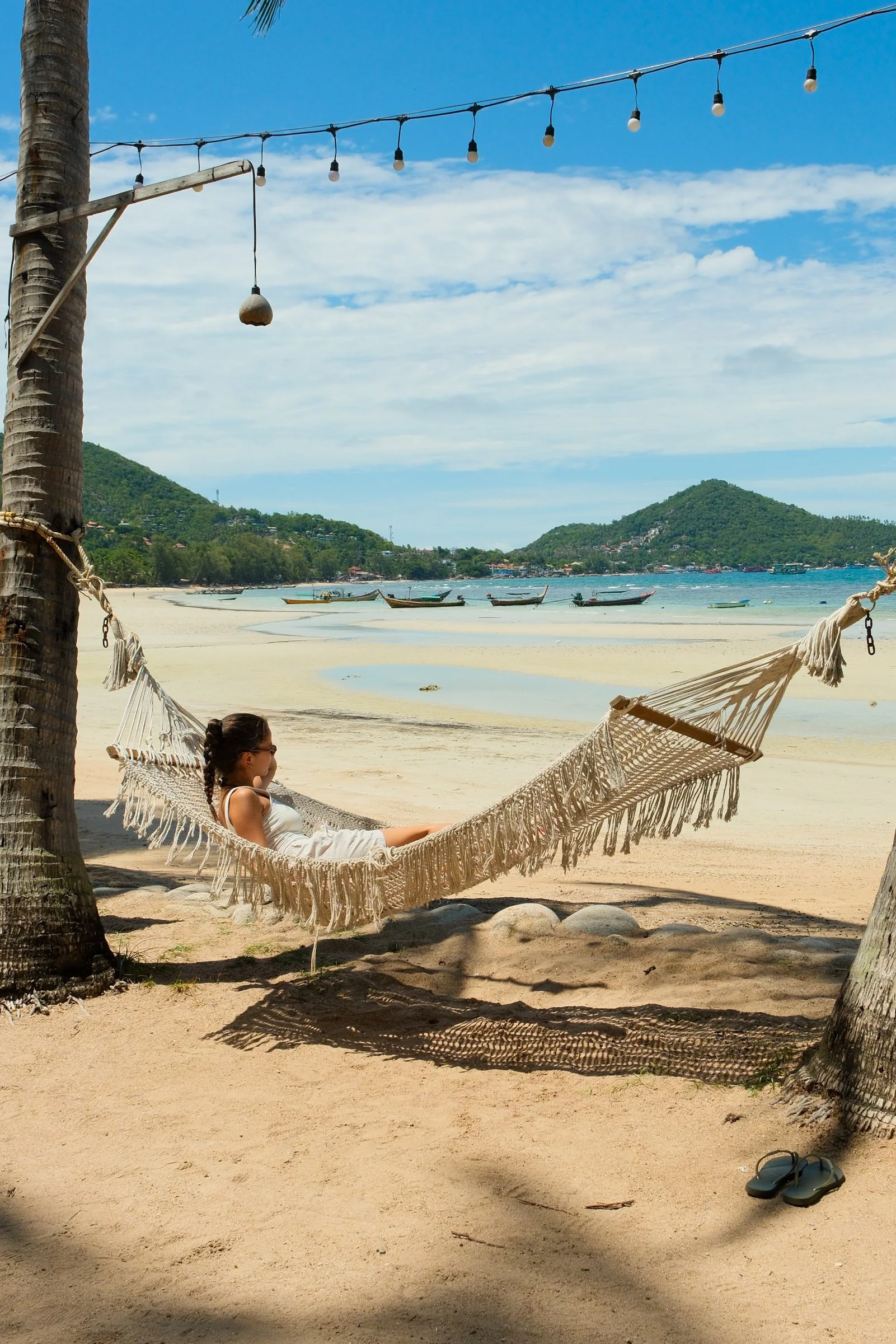 Guest relaxing in a hammock overlooking a sandy beach, turquoise bay, and traditional longtail boats