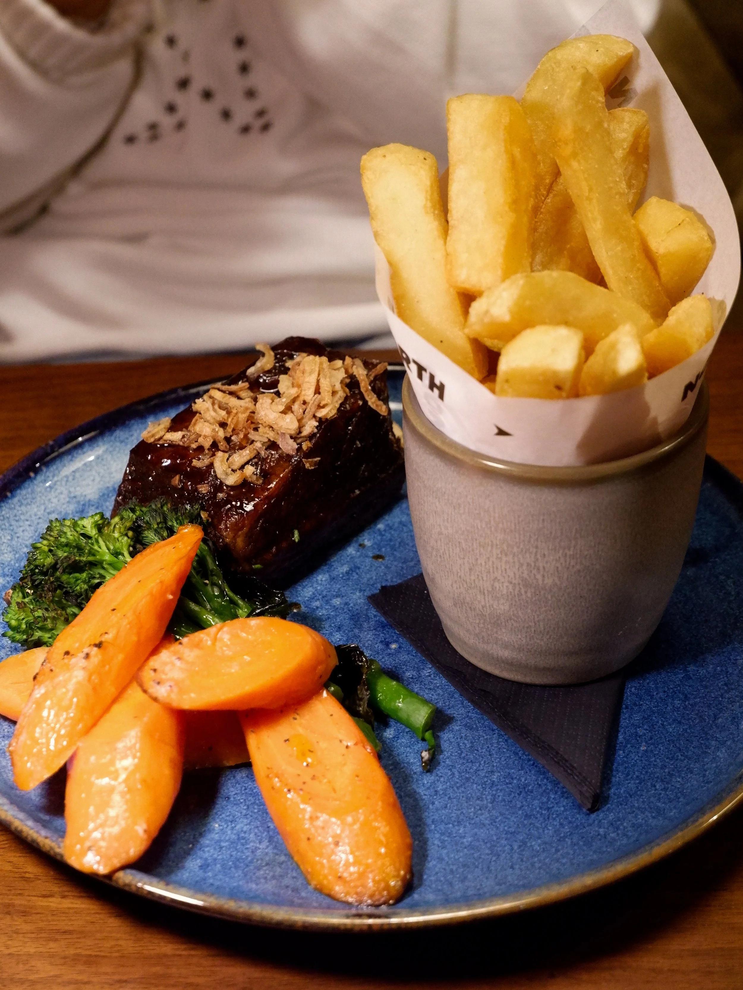 Glazed beef with crispy onions, roasted carrots, broccoli, and thick-cut chips on a blue ceramic plate