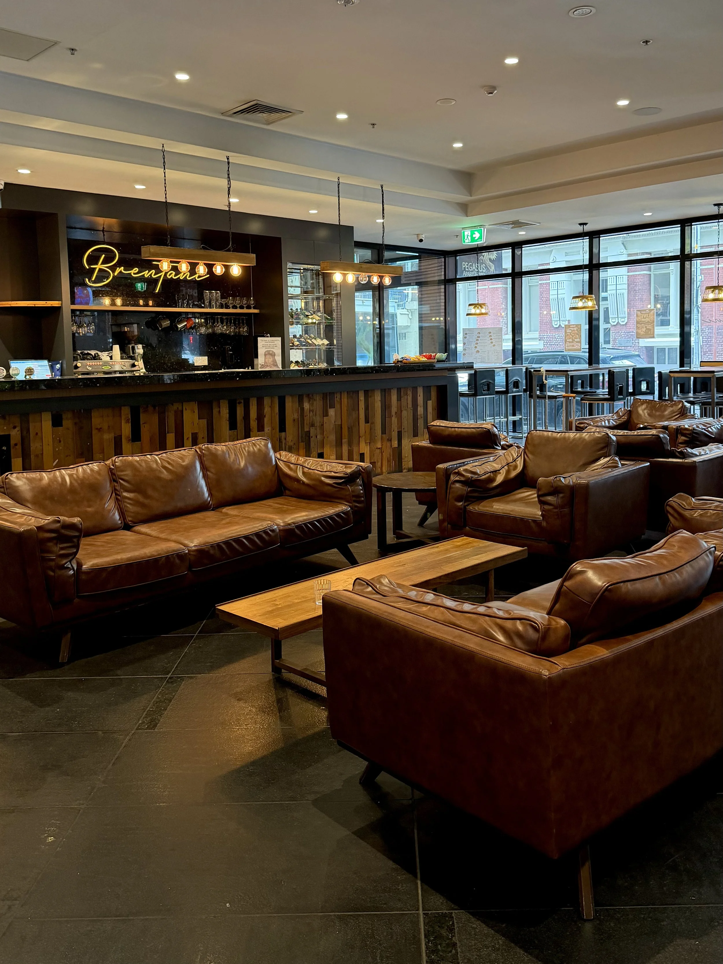 Comfortable hotel lounge with brown leather sofas, wooden coffee tables, and a bar overlooking the city street