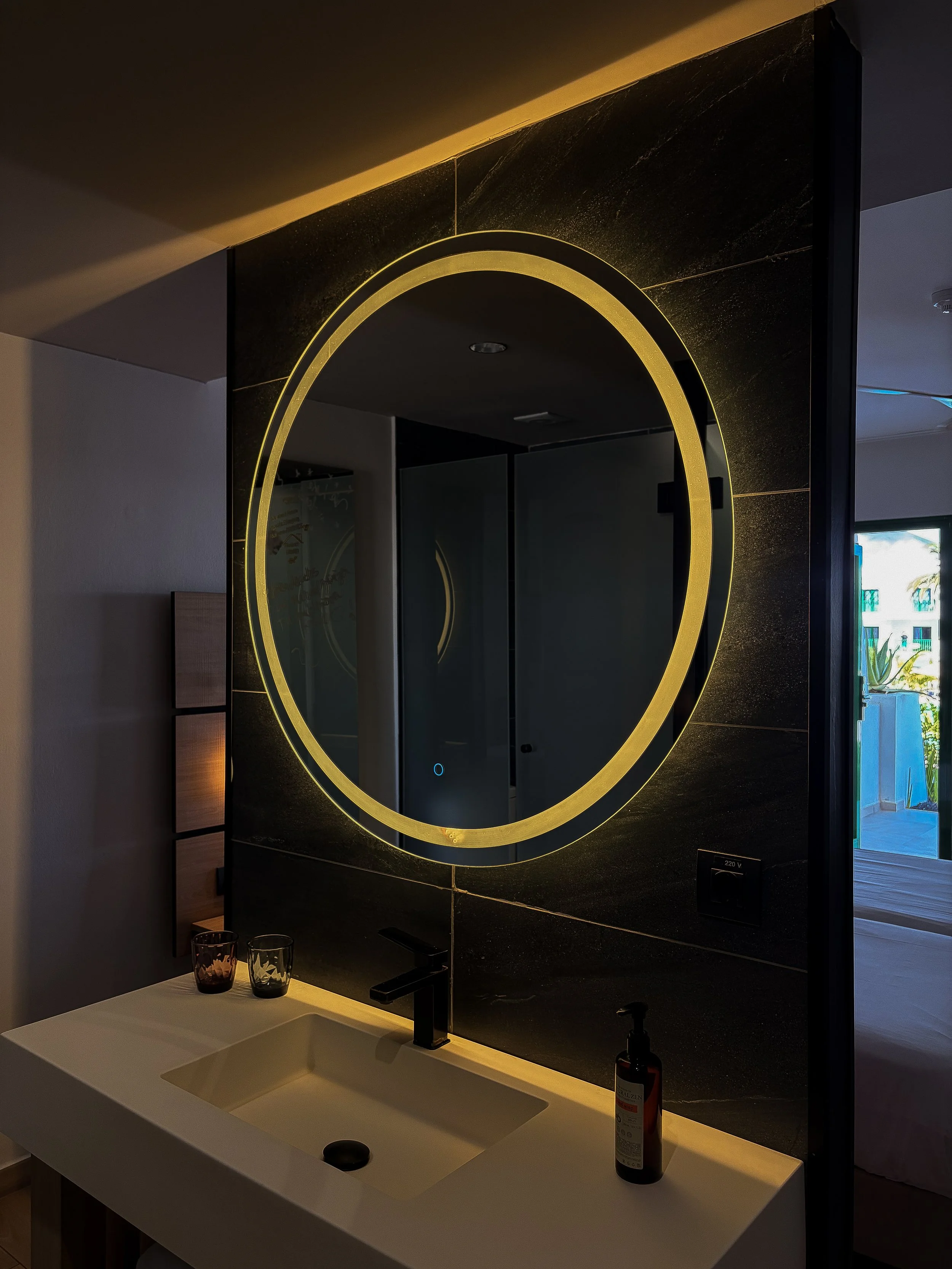 Modern bathroom vanity with a circular illuminated mirror, black stone tiles, and minimalist fixtures
