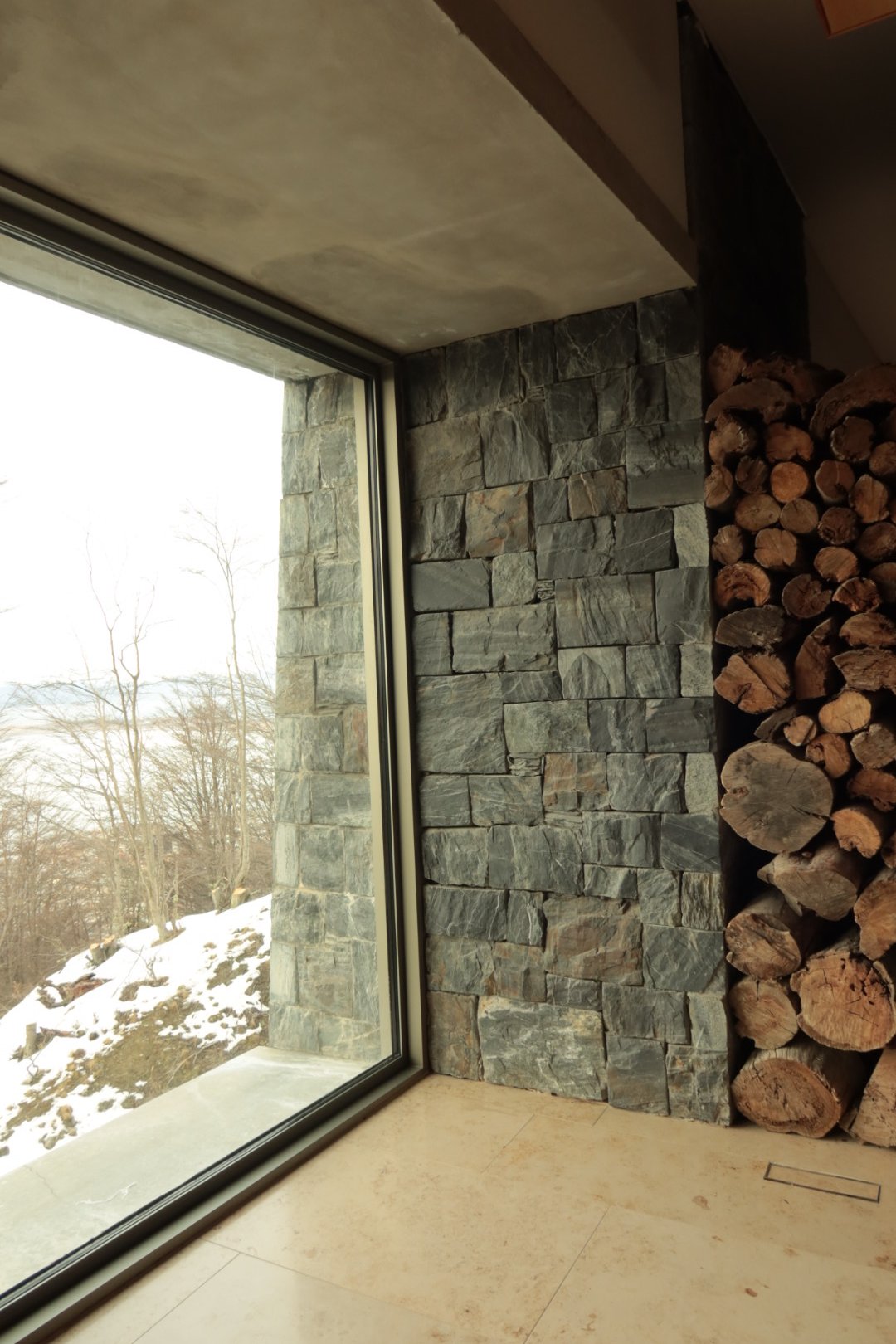 Floor-to-ceiling window overlooking snowy hills beside natural stone walls and stacked firewood