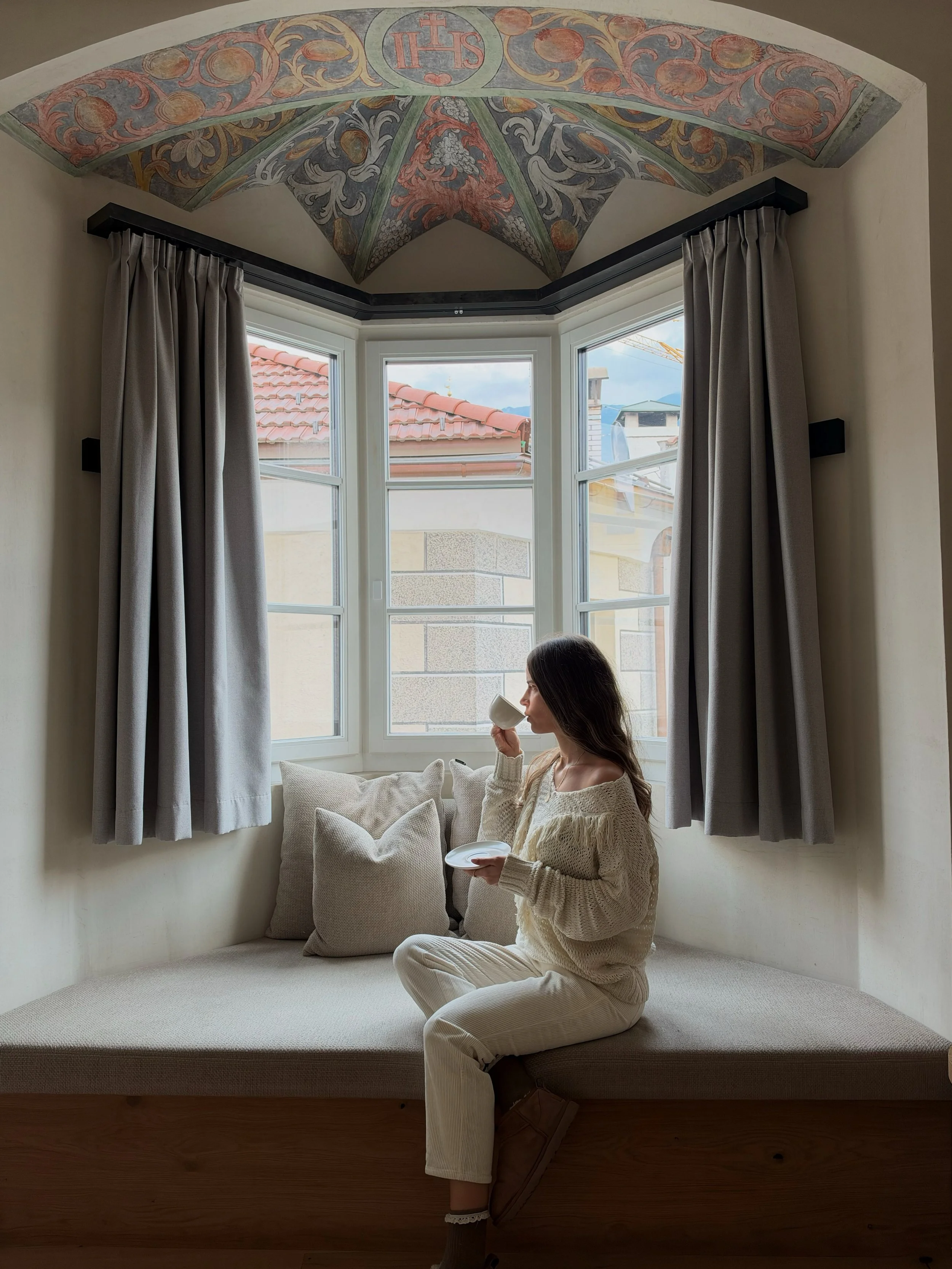 Guest enjoying a morning coffee beside a bay window with historic painted ceiling details and comfortable seating