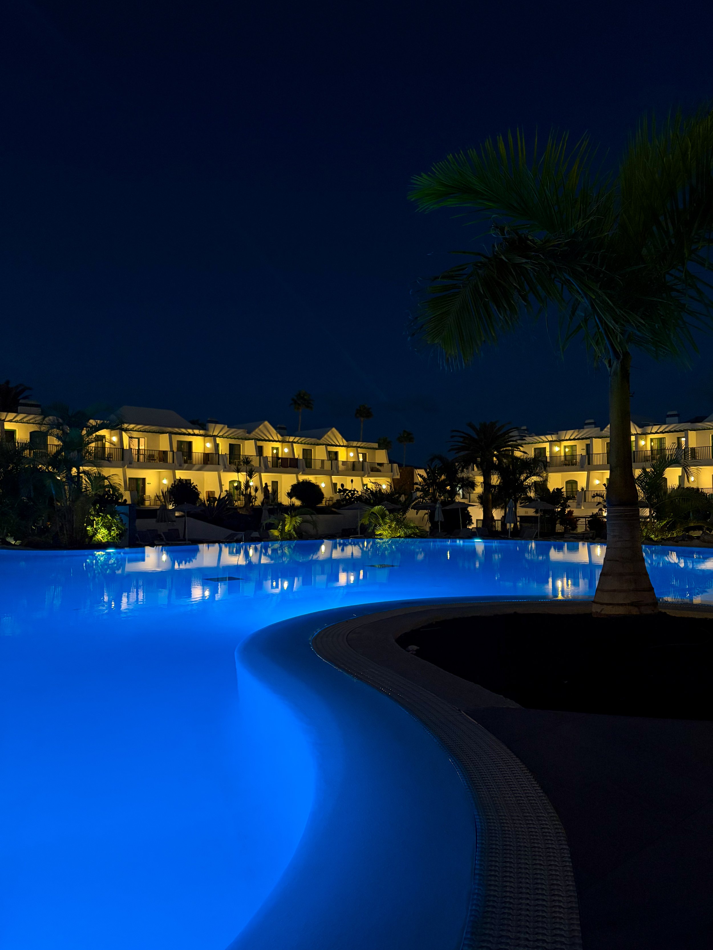 Illuminated swimming pool at night with glowing blue water, palm trees, and warmly lit guest room balconies