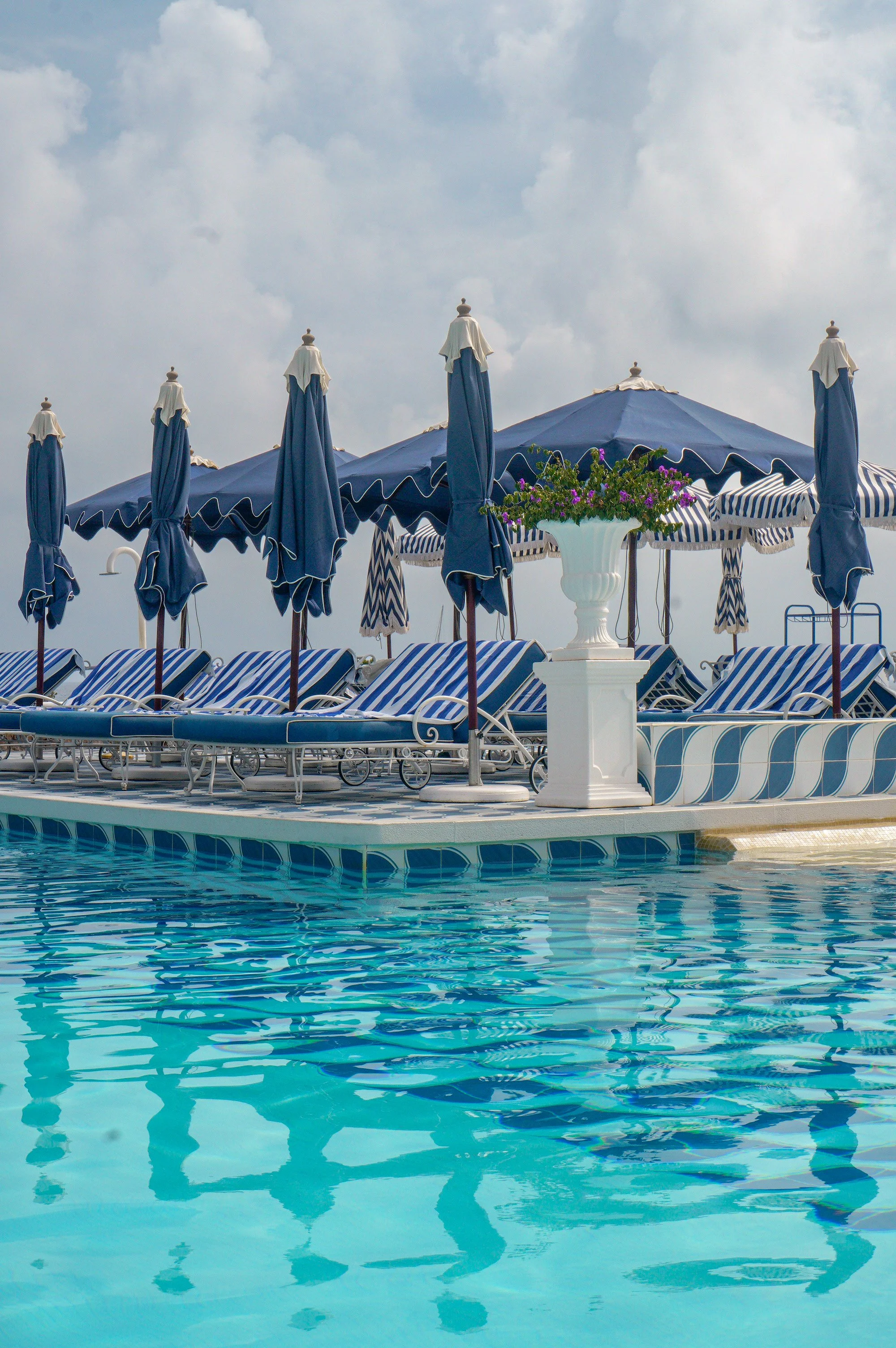 Outdoor swimming pool surrounded by blue and white striped sun loungers, matching parasols, and Mediterranean views