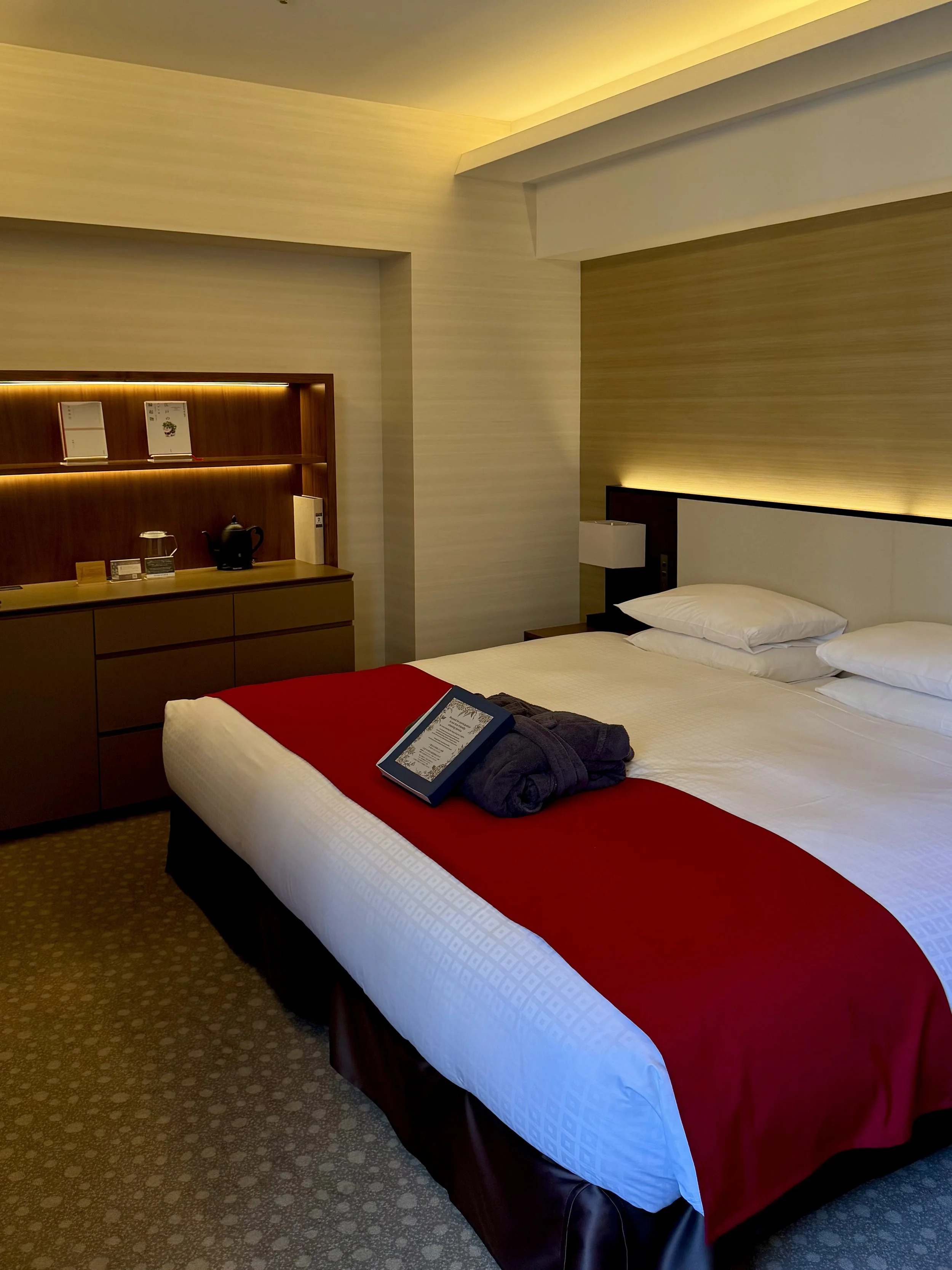 Warmly lit guest room with a double bed, red bed runner, tea station, and robes prepared for the stay