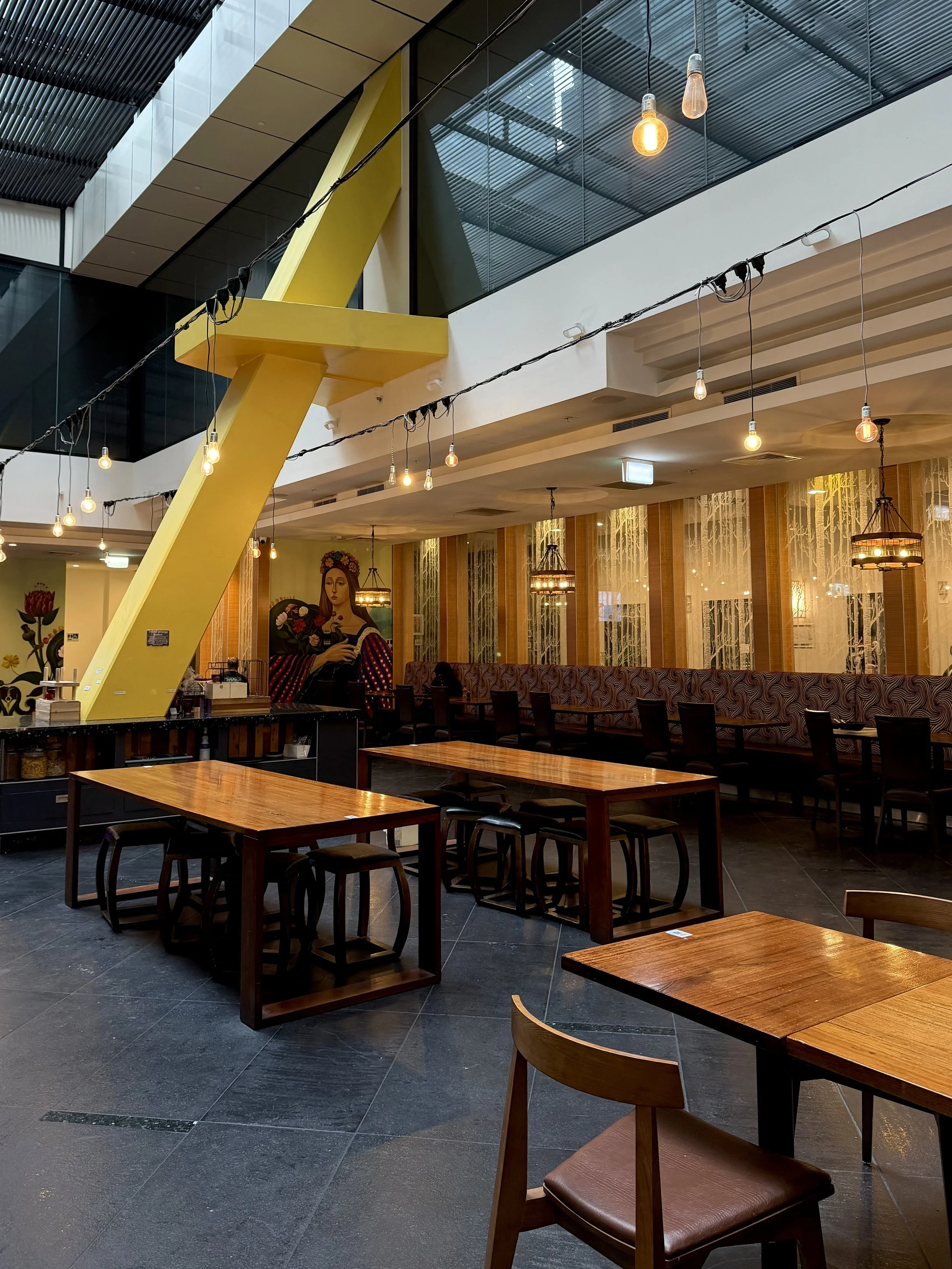 Spacious hotel dining area featuring wooden tables, modern lighting, a high ceiling, and contemporary architectural details