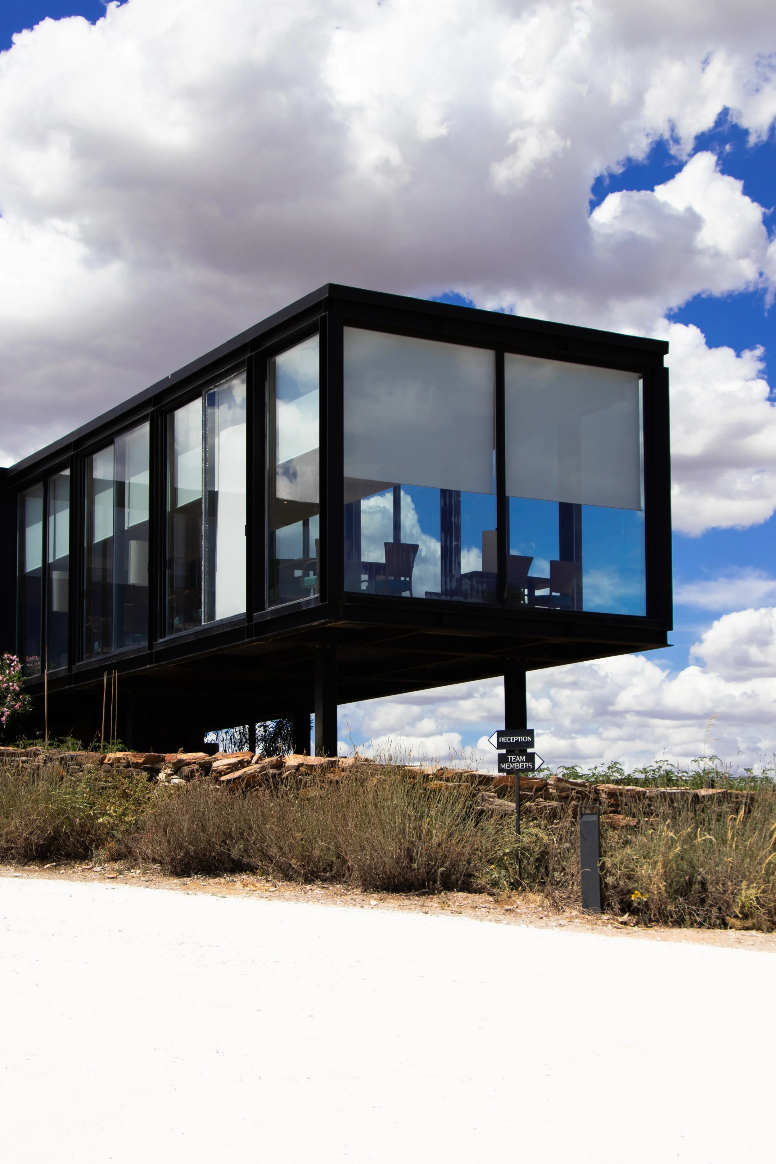Modern glass reception pavilion elevated above the landscape, reflecting blue skies and countryside views in minimalist architecture, Land of Alandroal – Alandroal, Portugal.