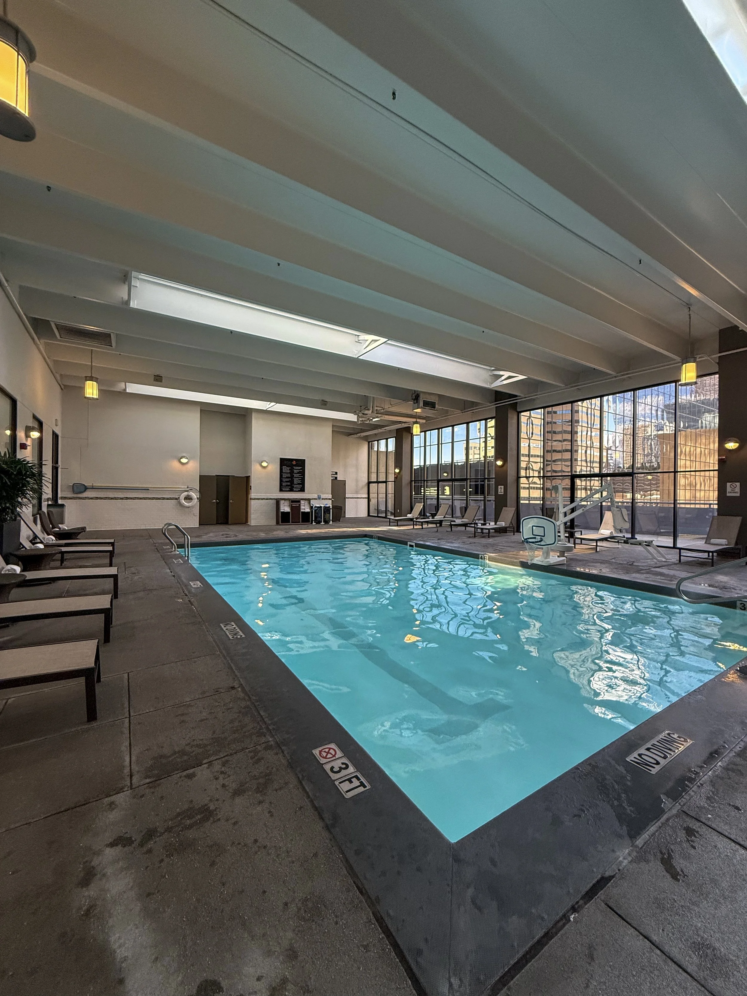 Indoor swimming pool surrounded by loungers and large windows overlooking downtown