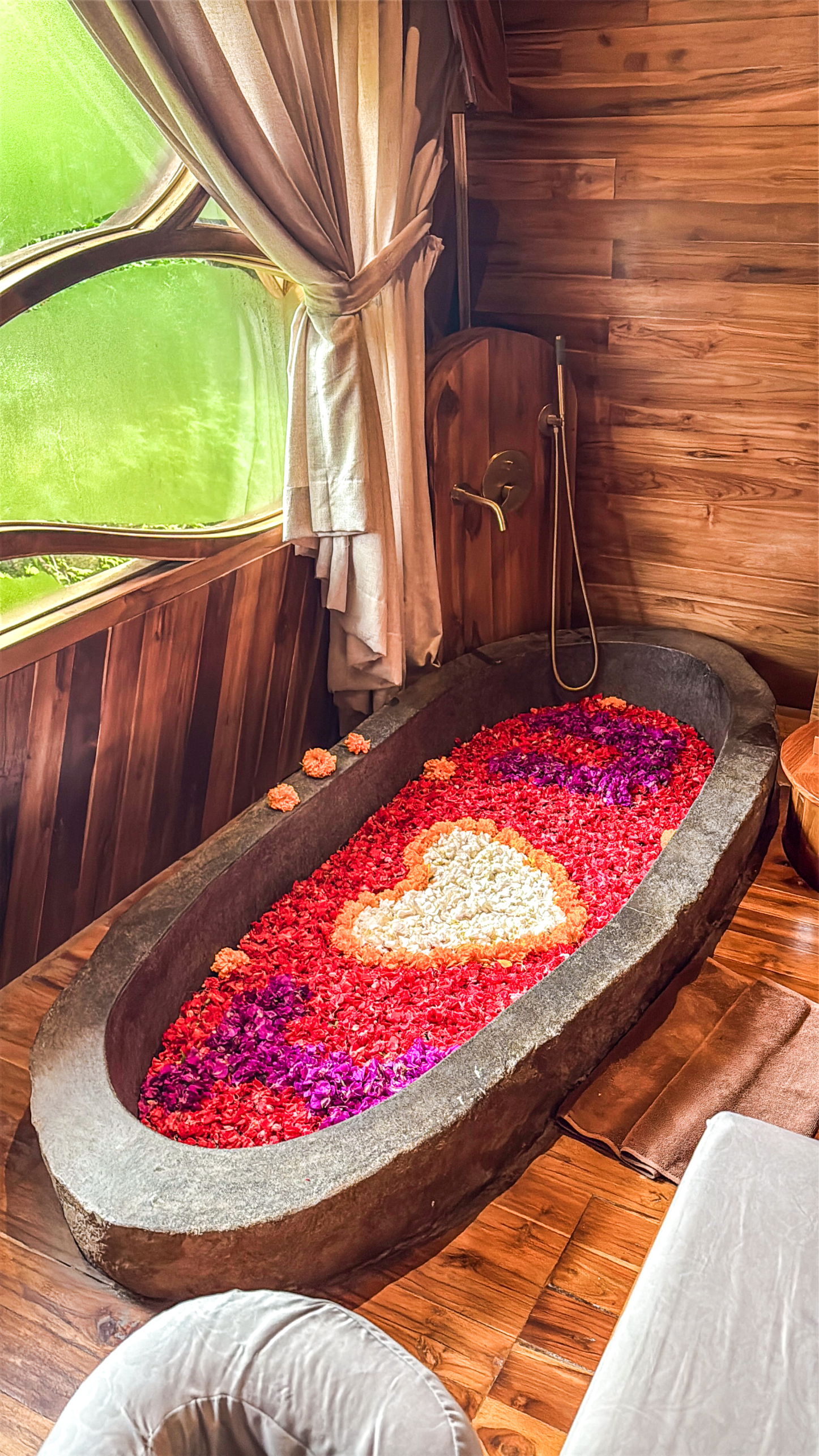 A handcrafted stone bathtub filled with flower petals brings together natural materials, warm wooden interiors, and a relaxing wellness experience