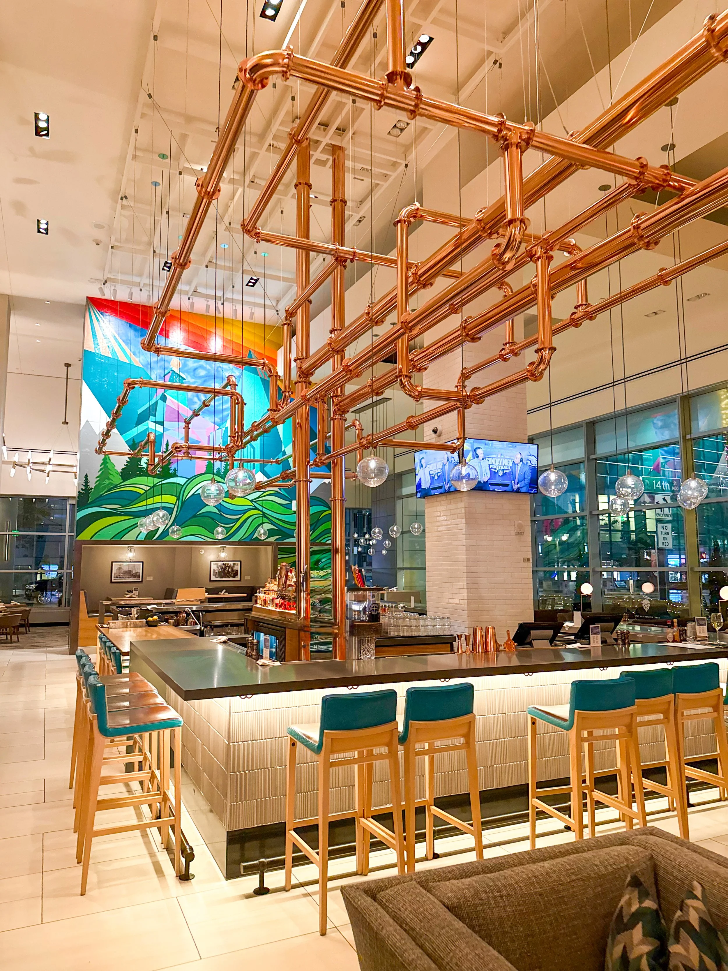 Modern Assembly Hall Bar + Market featuring turquoise bar stools, copper ceiling fixtures, and colorful wall art
