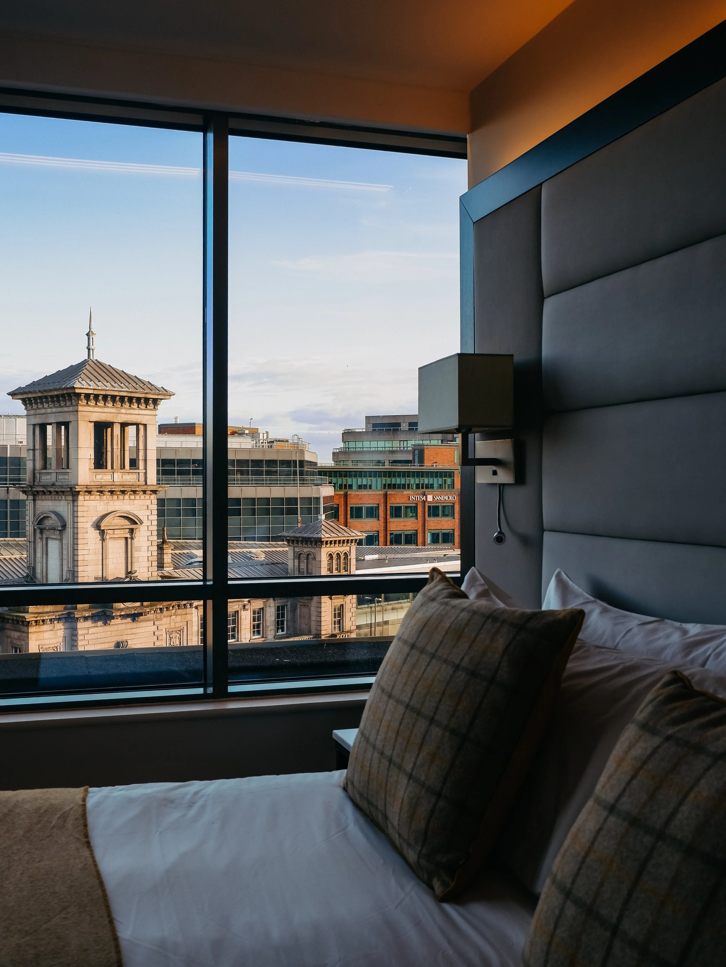 Comfortable hotel bed with a padded headboard and large windows framing Dublin's historic architecture