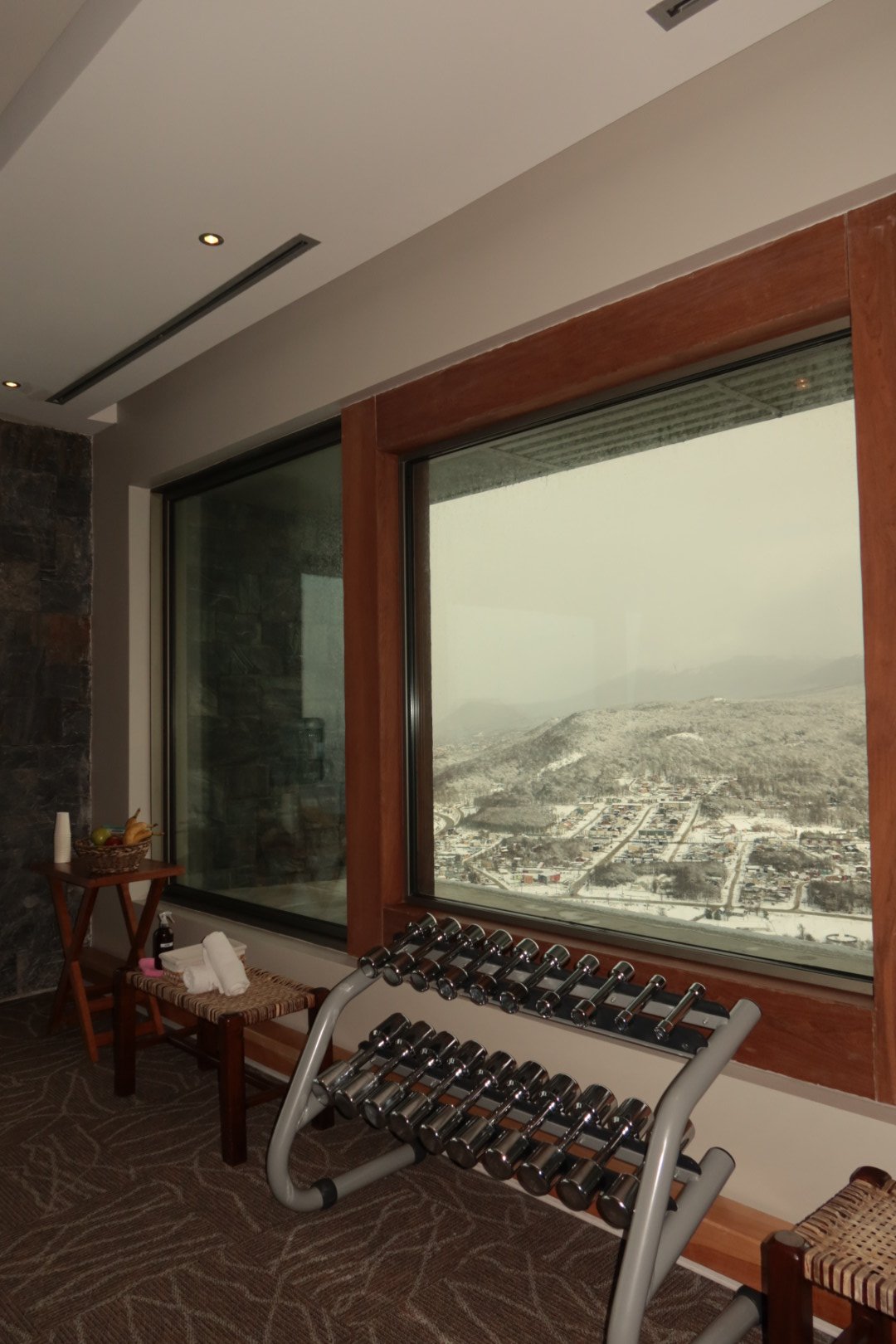 Fitness area with dumbbells positioned beside panoramic windows overlooking snow-covered mountains and Ushuaia
