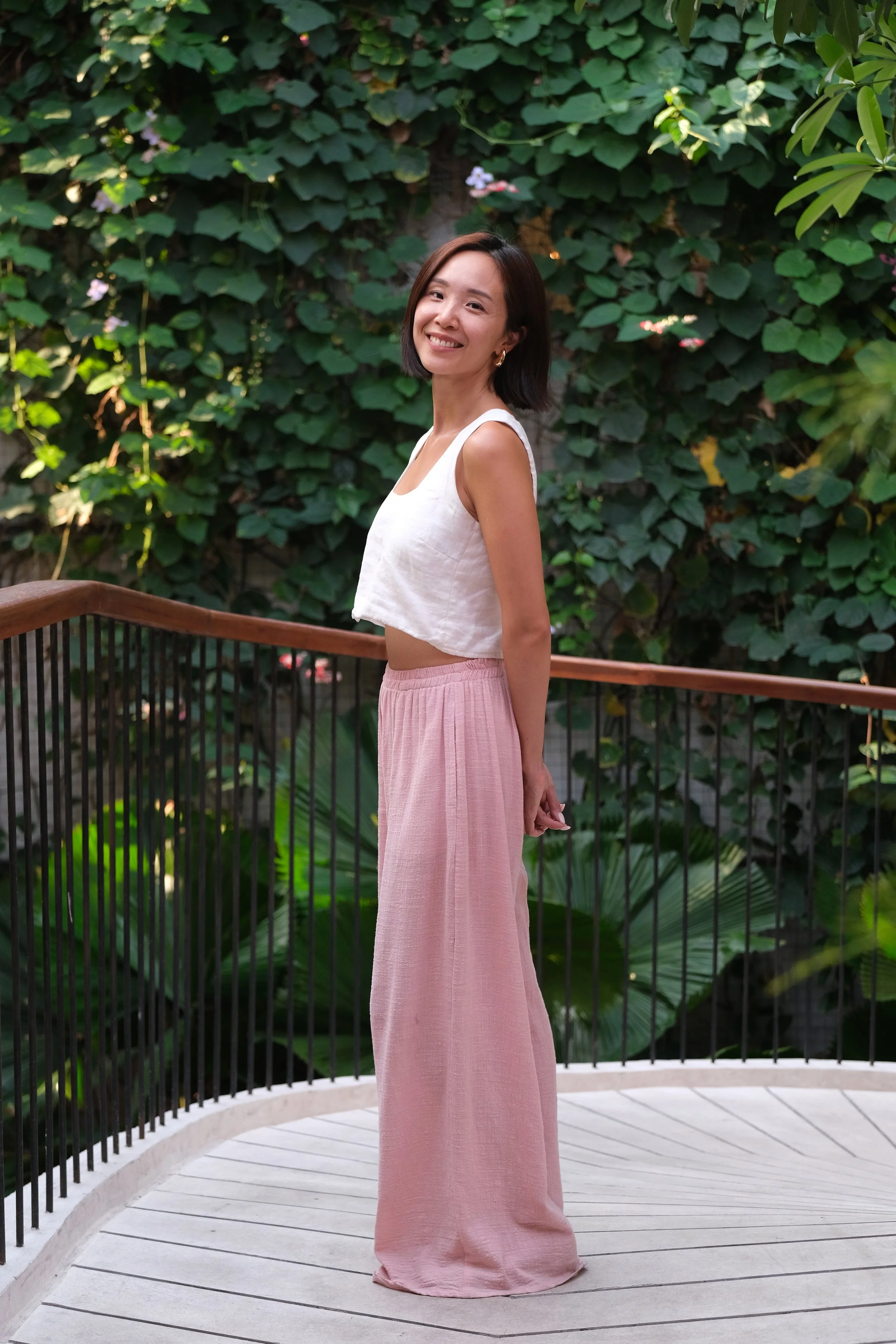 A woman is standing on a curved wooden deck surrounded by lush green foliage, smiling at the camera. She is wearing a white sleeveless crop top and pink wide-leg pants.