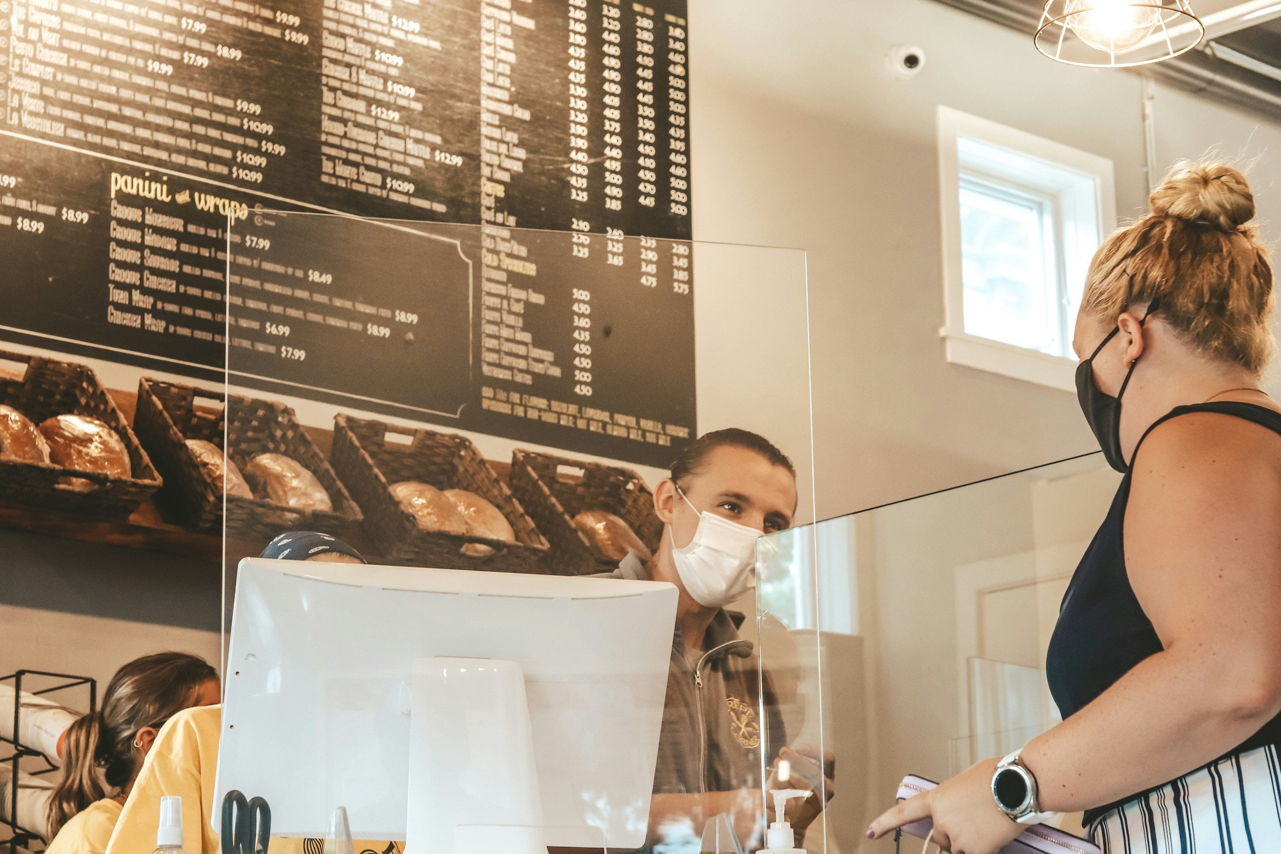 A woman wearing a black face mask and black sleeveless shirt talking to a cashier behind a transparent protective barrier in a cafe. The cashier, also wearing a face mask, is looking at her. The woman has a striped skirt and a smartwatch. In the background, a large menu board is visible on the wall with various food and drink options.