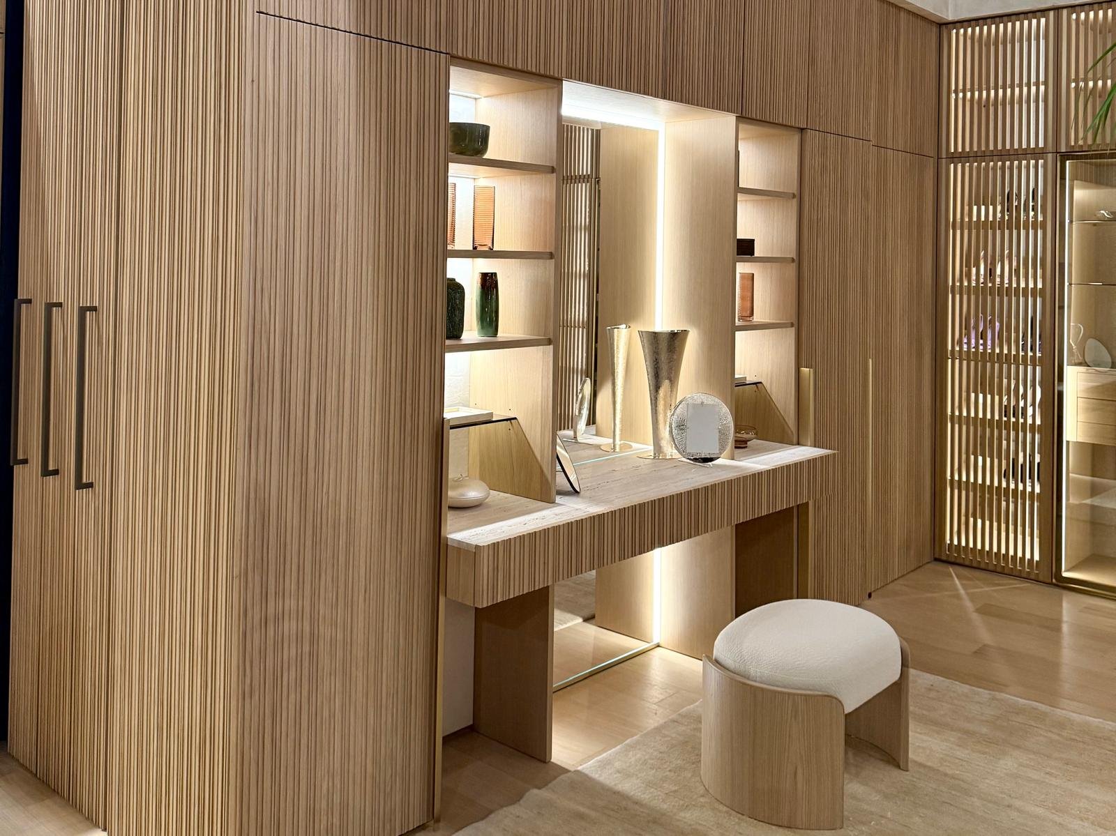 Interno di una stanza con mobili in legno chiaro, scaffali con vasi decorativi e un piccolo tavolo con oggetti decorativi. Presenza di una seduta imbottita. L'ambiente è luminoso e moderno.