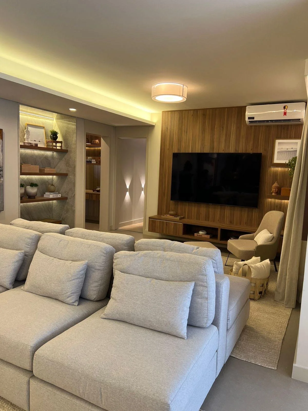 Salotto accogliente con un'illuminazione calda e un grande divano grigio chiaro. La parete in legno e la TV a parete completano l'aspetto moderno e funzionale del soggiorno.