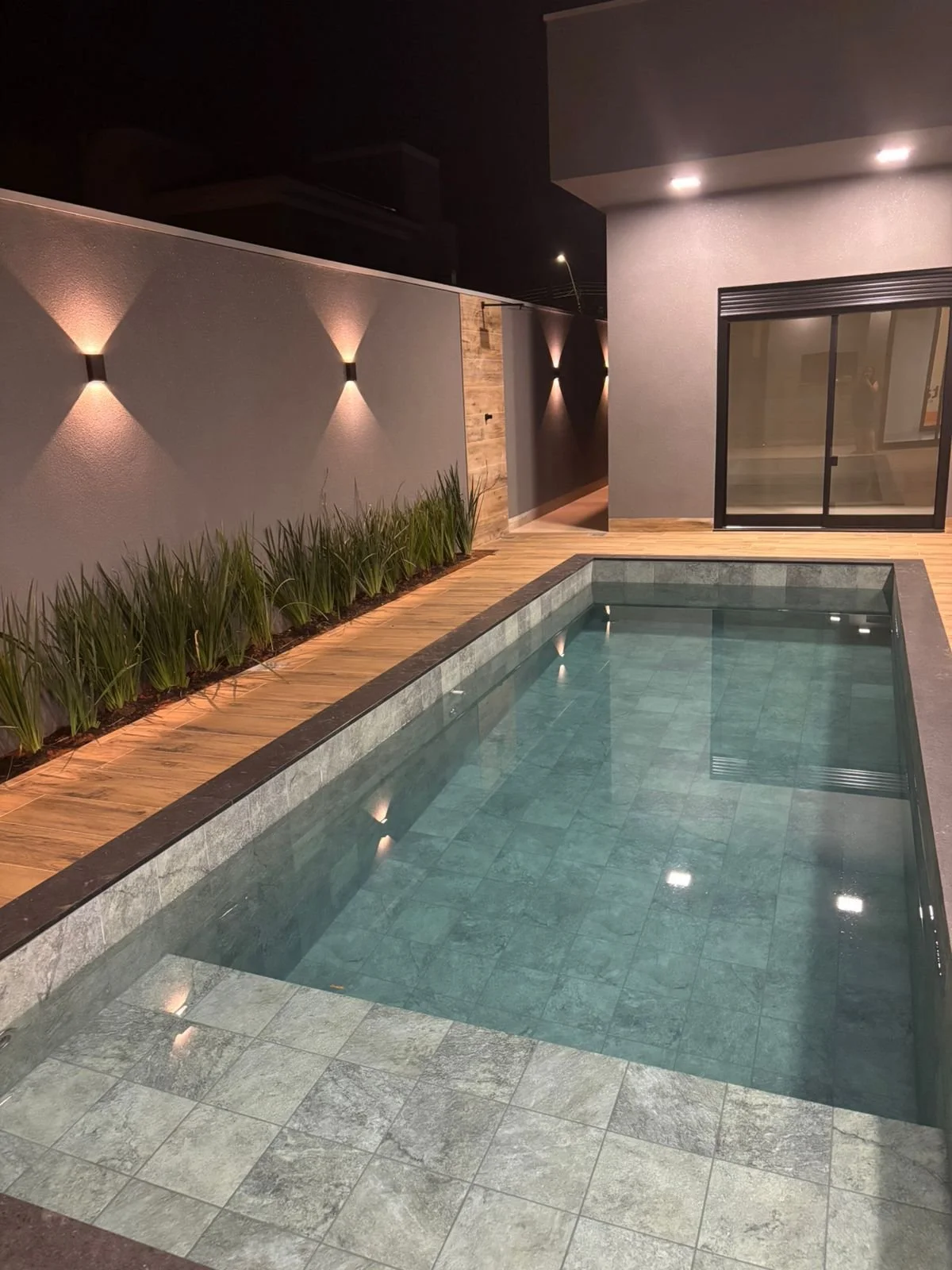 Il riflesso dell'illuminazione notturna sulla piscina lineare definisce il carattere moderno di questo spazio esterno. La fioriera laterale integrata completa la visione d'insieme, creando un ambiente outdoor di alto livello. 