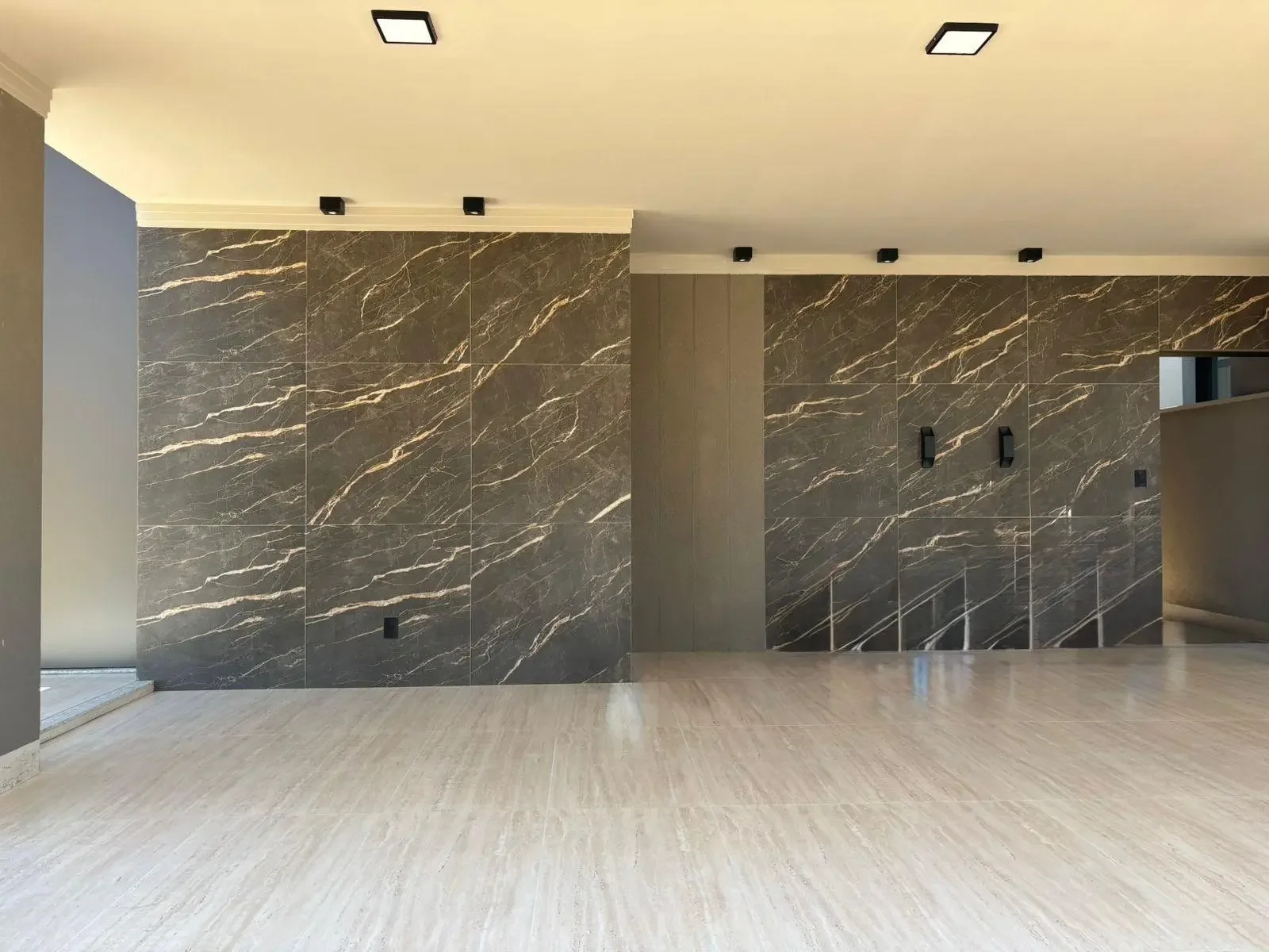 Parete interamente rivestita con grandi lastre di ceramica effetto marmo marrone, arricchite da venature chiare e dinamiche. L'illuminazione è curata con faretti a soffitto minimalisti e applique bidirezionali nere che creano giochi di luce radente s