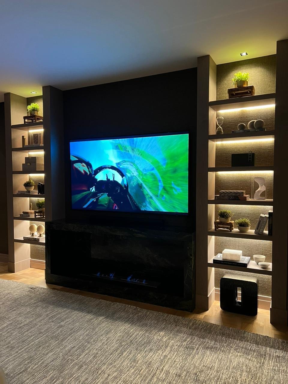 Grande TV a parete e mensole illuminate. L'uso della luce calda e degli spazi organizzati offre eleganza e comfort.