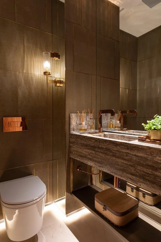 Bagno di servizio scenografico con rivestimento bronzo, luce calda e dettagli metallici coordinati.