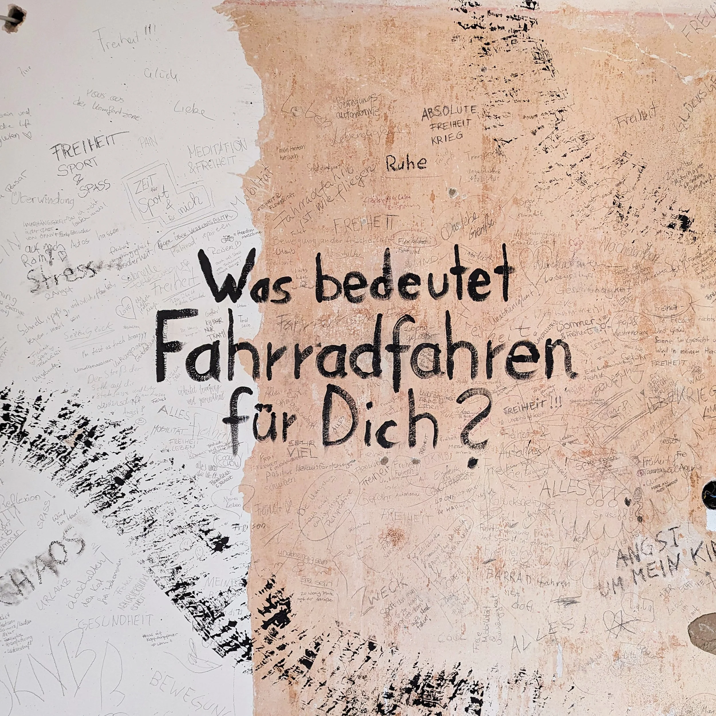 A wall with many handwritten notes and messages in German surrounding the large black text, 'Was bedeutet Fahrradfahren für Dich?' which translates to 'What does cycling mean to you?' in English.